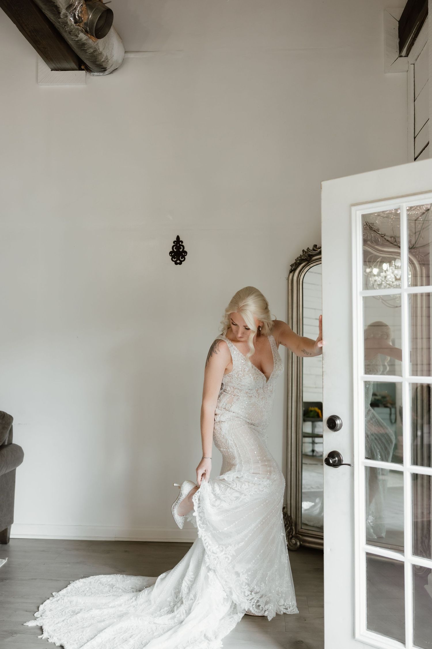 Blonde bride in fitted lace wedding gown with train standing by French doors in bridal suite.