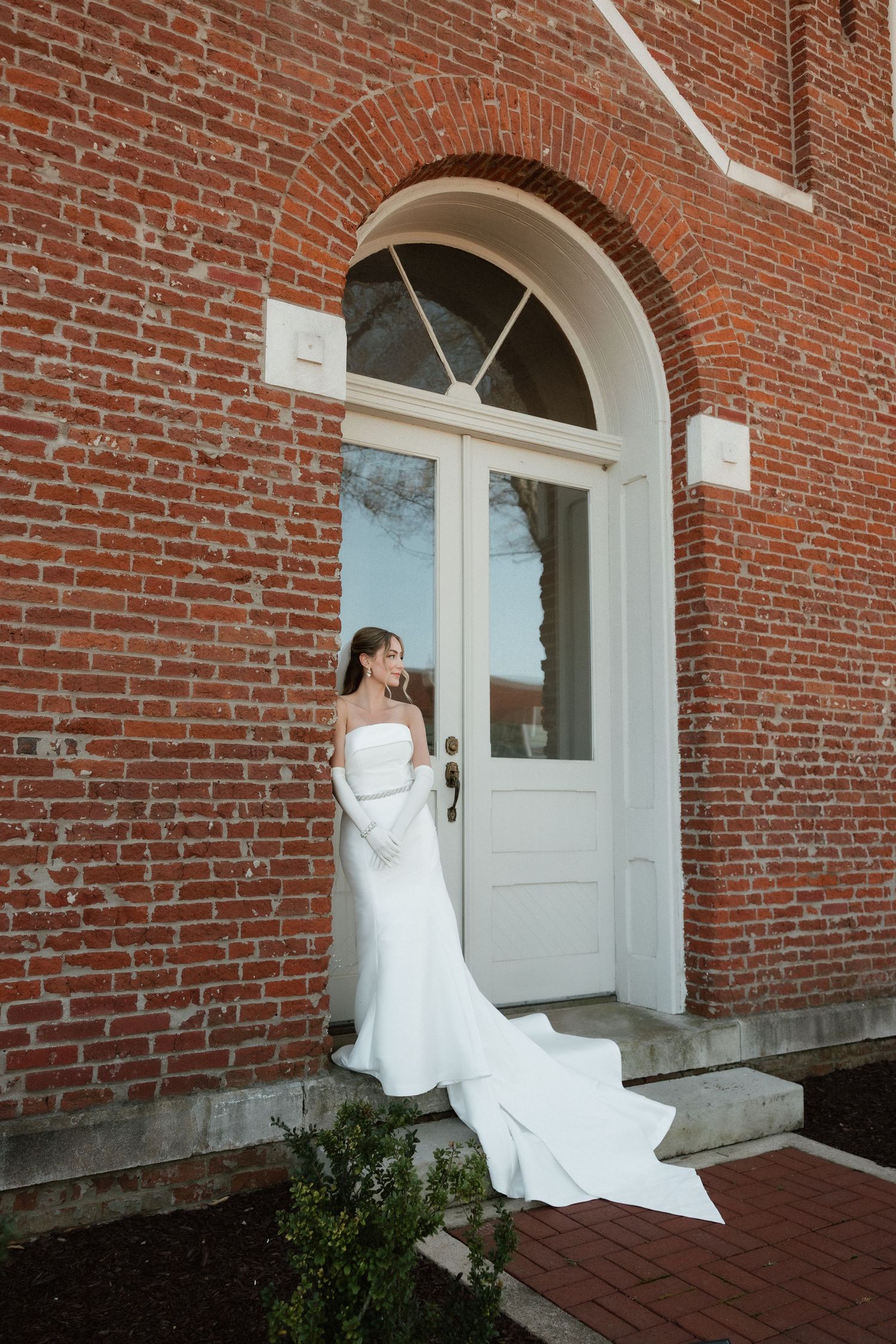 Bride in strapless white gown with long train stands before an arched door of a brick building.