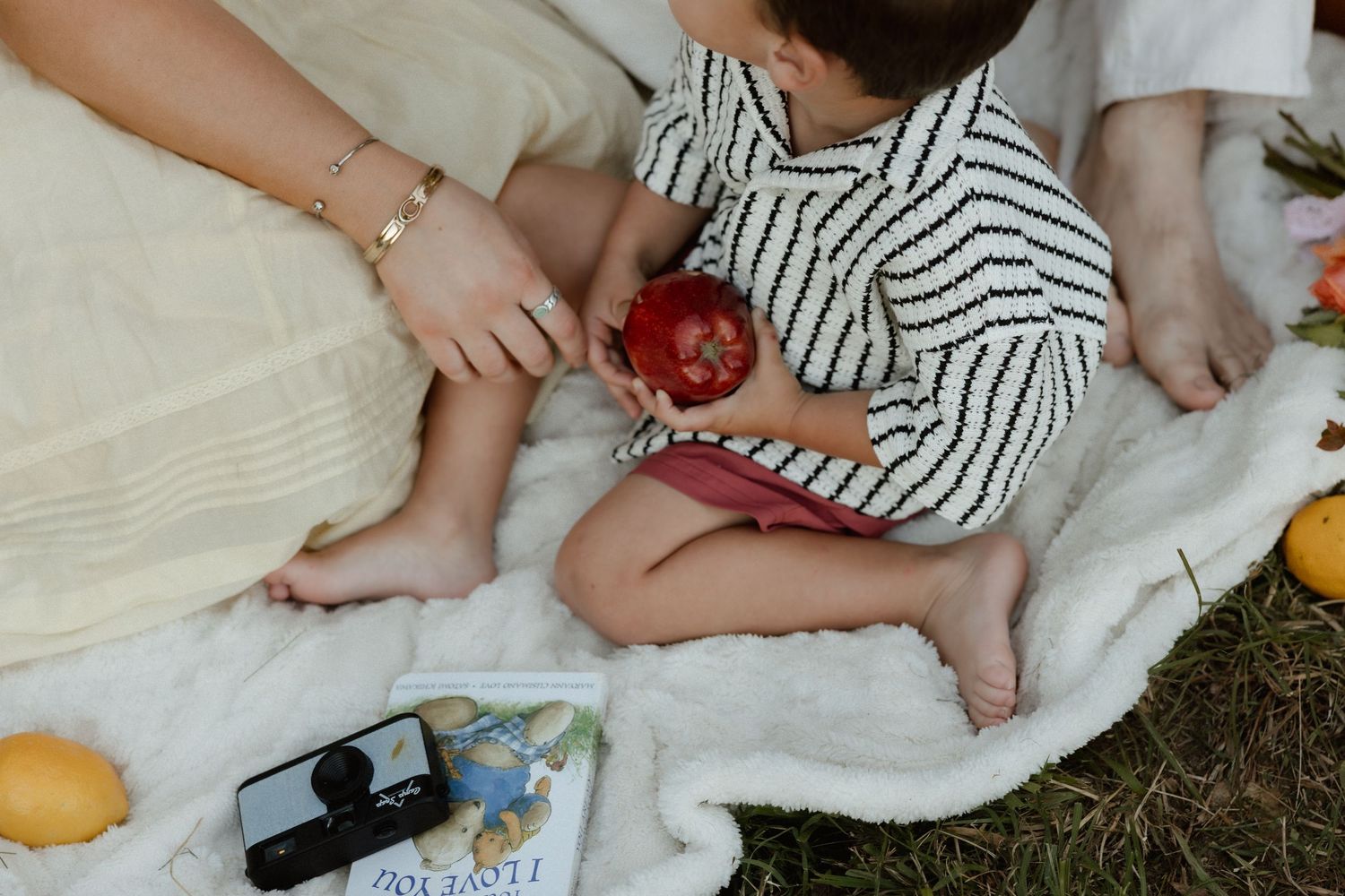 Baby in striped outfit sitting on white blanket holding a red apple during an outdoor picnic.