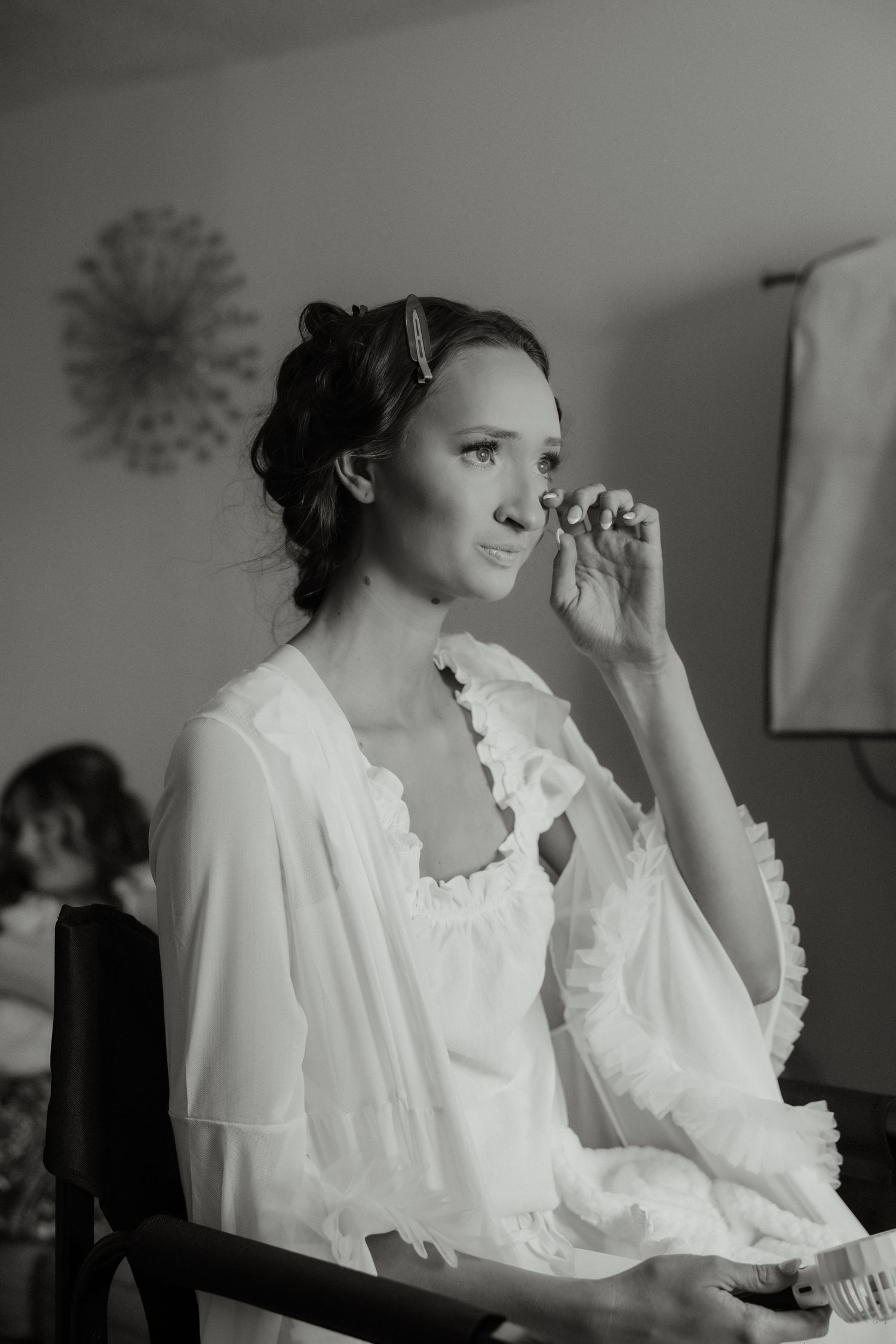A black and white photo of a bride wiping a tear during wedding preparations in a bright room.