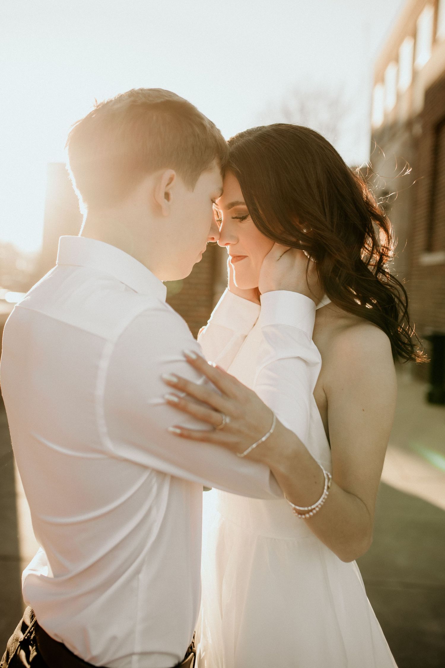 Couple embracing in golden sunset light, woman in white dress touching mans face in romantic outdoor portrait.