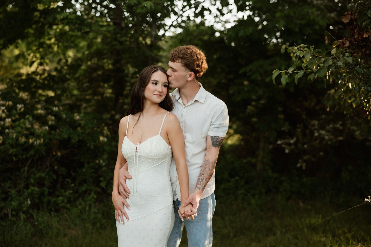 A couple stands together in golden hour light as the man kisses the womans cheek outdoors.