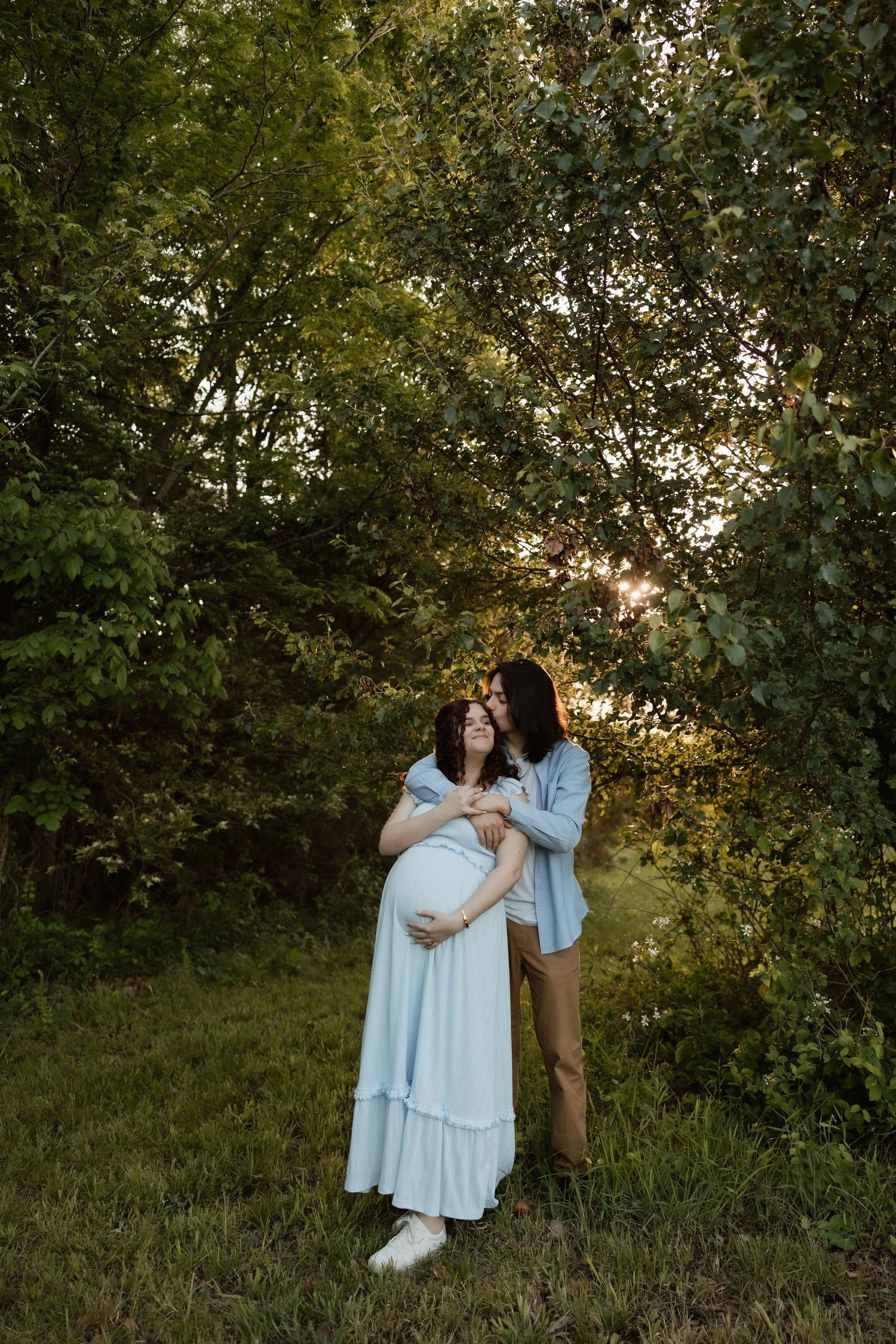 Pregnant couple embracing at sunset in lush green forest setting.