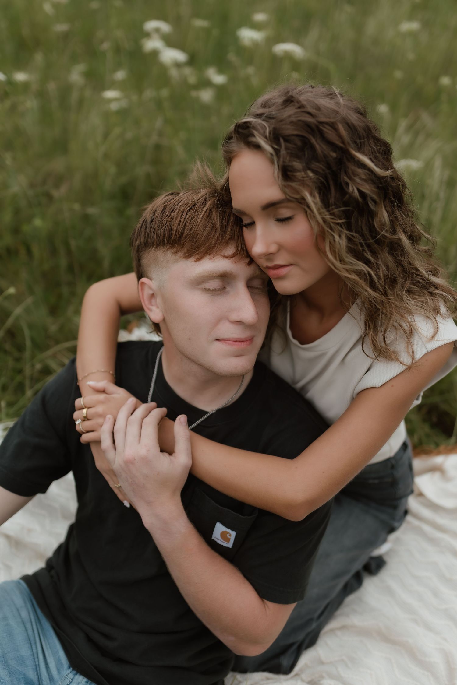 Woman with curly hair embracing man from behind as they sit on blanket in lush green meadow.