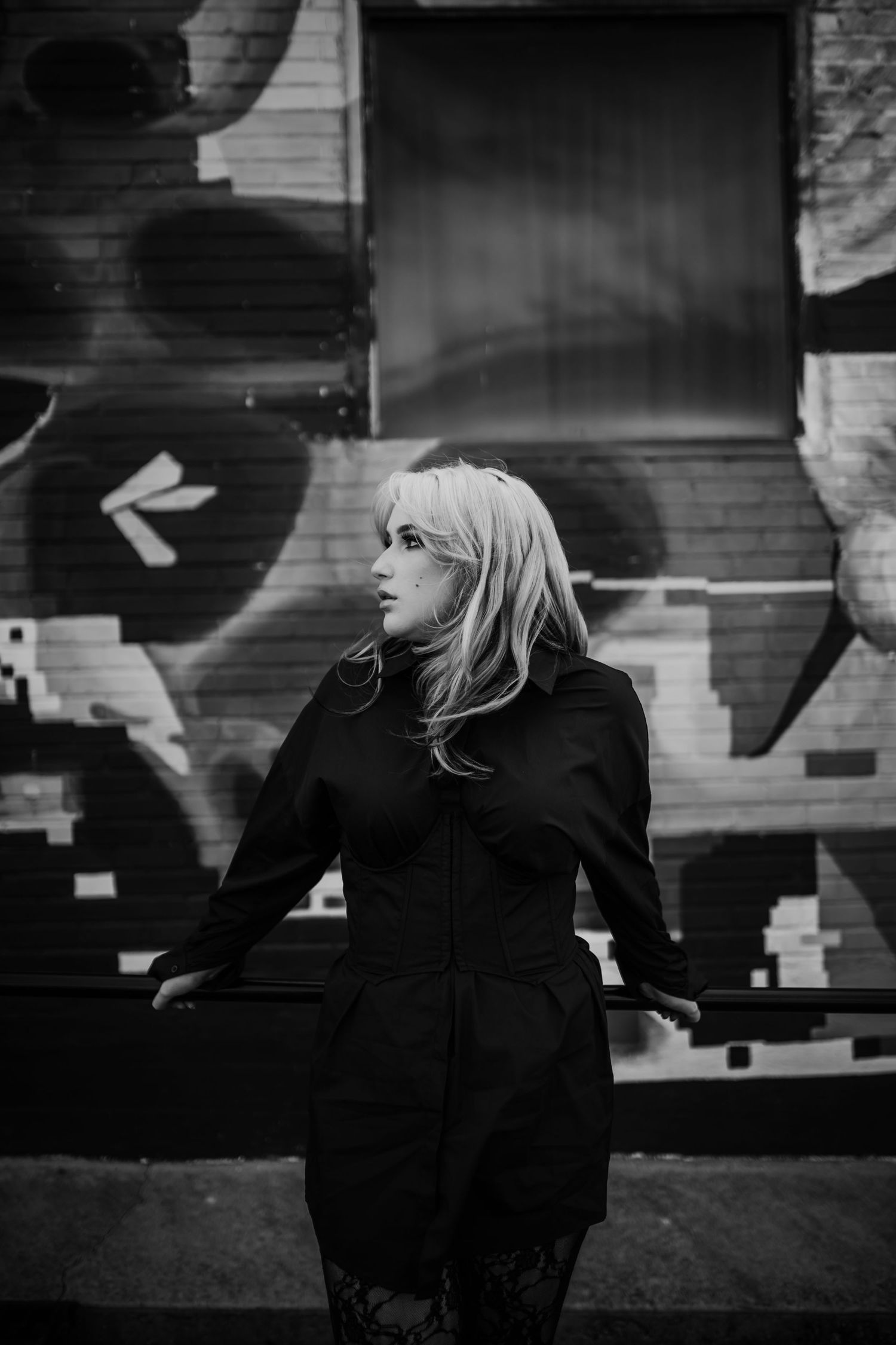 Black and white photo of blonde woman leaning on rail by graffiti wall.