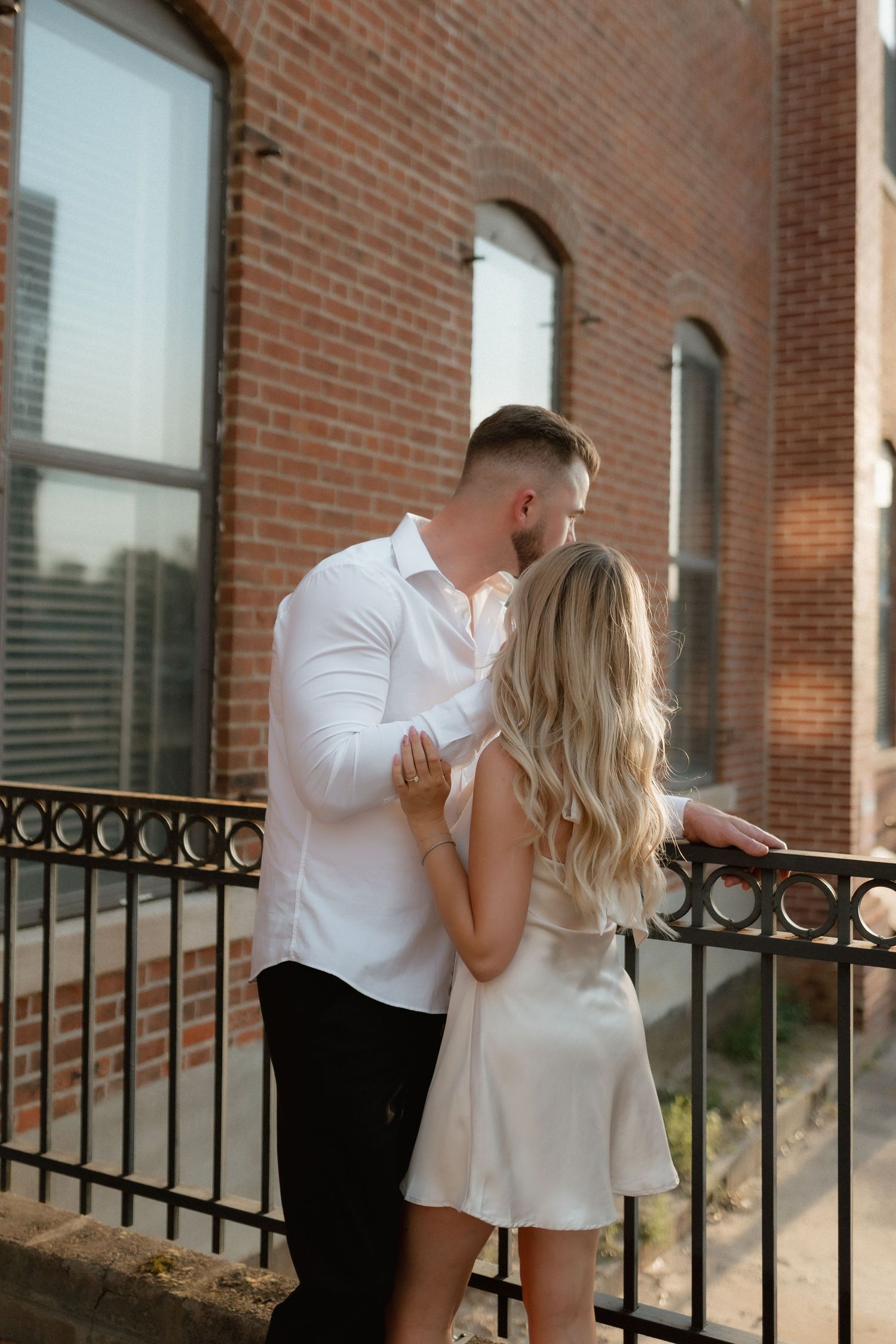 Couple sharing kiss on iron railing balcony outside historic red brick building in warm sunlight.