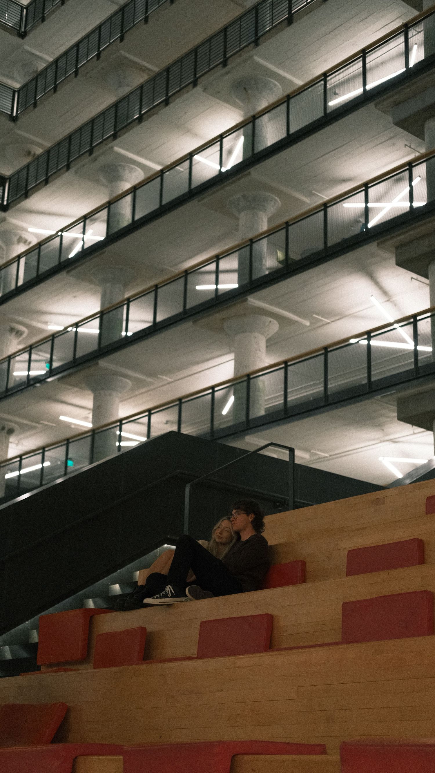 Person sitting alone on auditorium steps beneath multi-level illuminated building interior.
