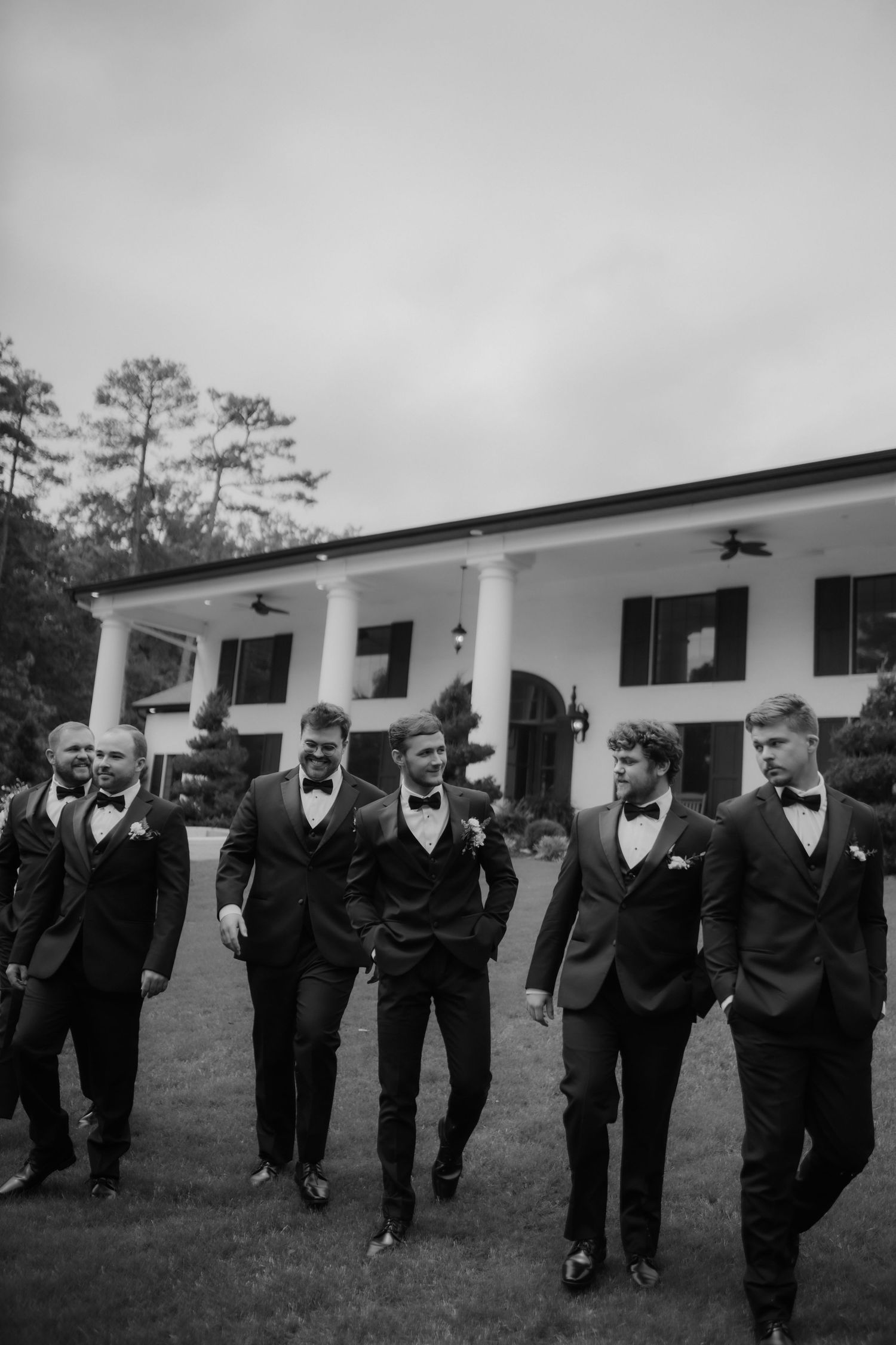 Groom and groomsmen in black tuxedos walking together outside elegant venue.
