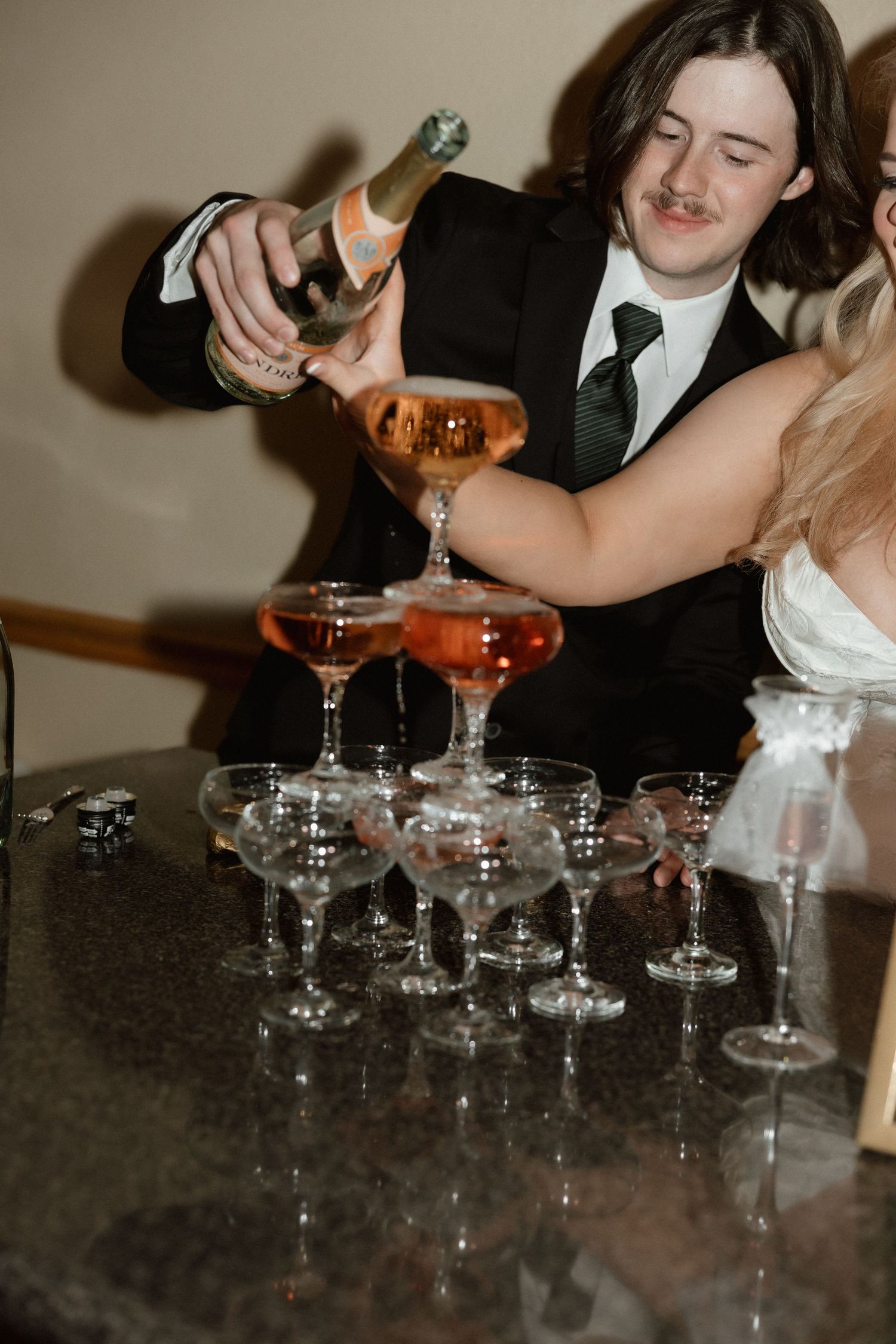 Groom pouring champagne into elegant coupe glass tower at wedding reception.
