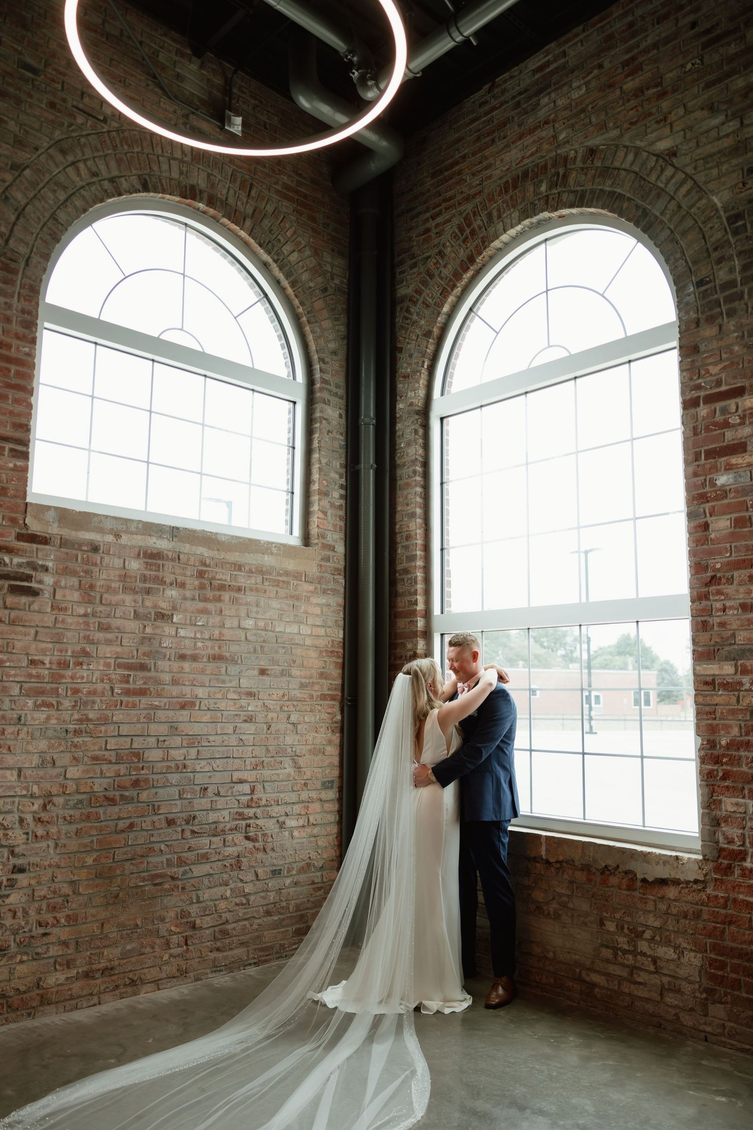 Bride and groom embrace near arched windows in rustic brick venue with dramatic cathedral veil flowing down.