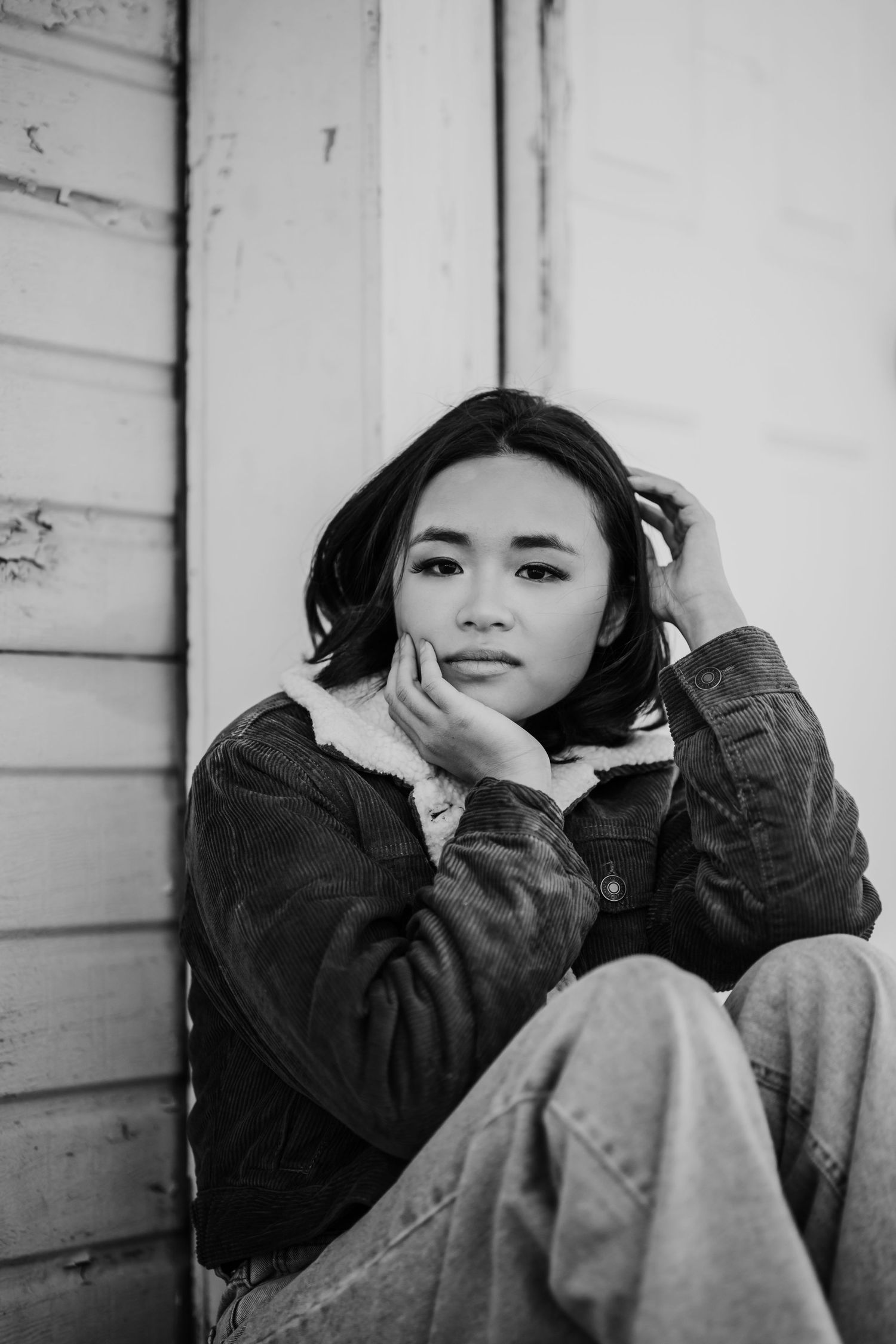 Black and white portrait of young Asian woman in denim jacket sitting against white wooden wall.