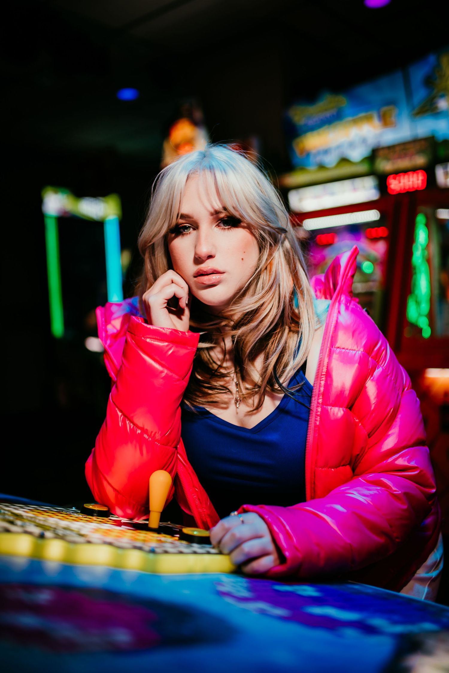 Blonde teen in pink puffer jacket posing at neon-lit arcade game.