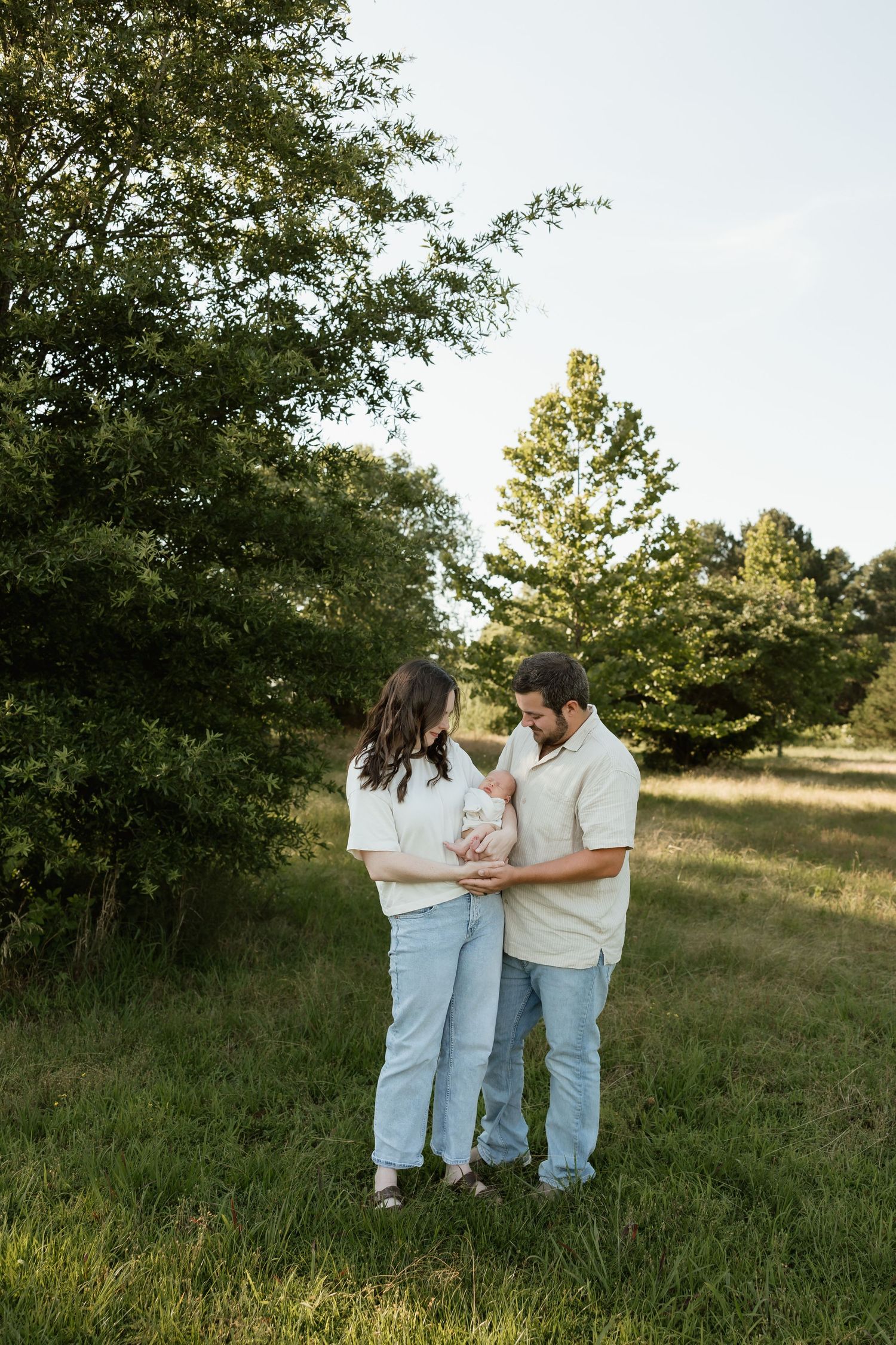 Couple standing in green field holding newborn baby, surrounded by lush trees during golden hour.