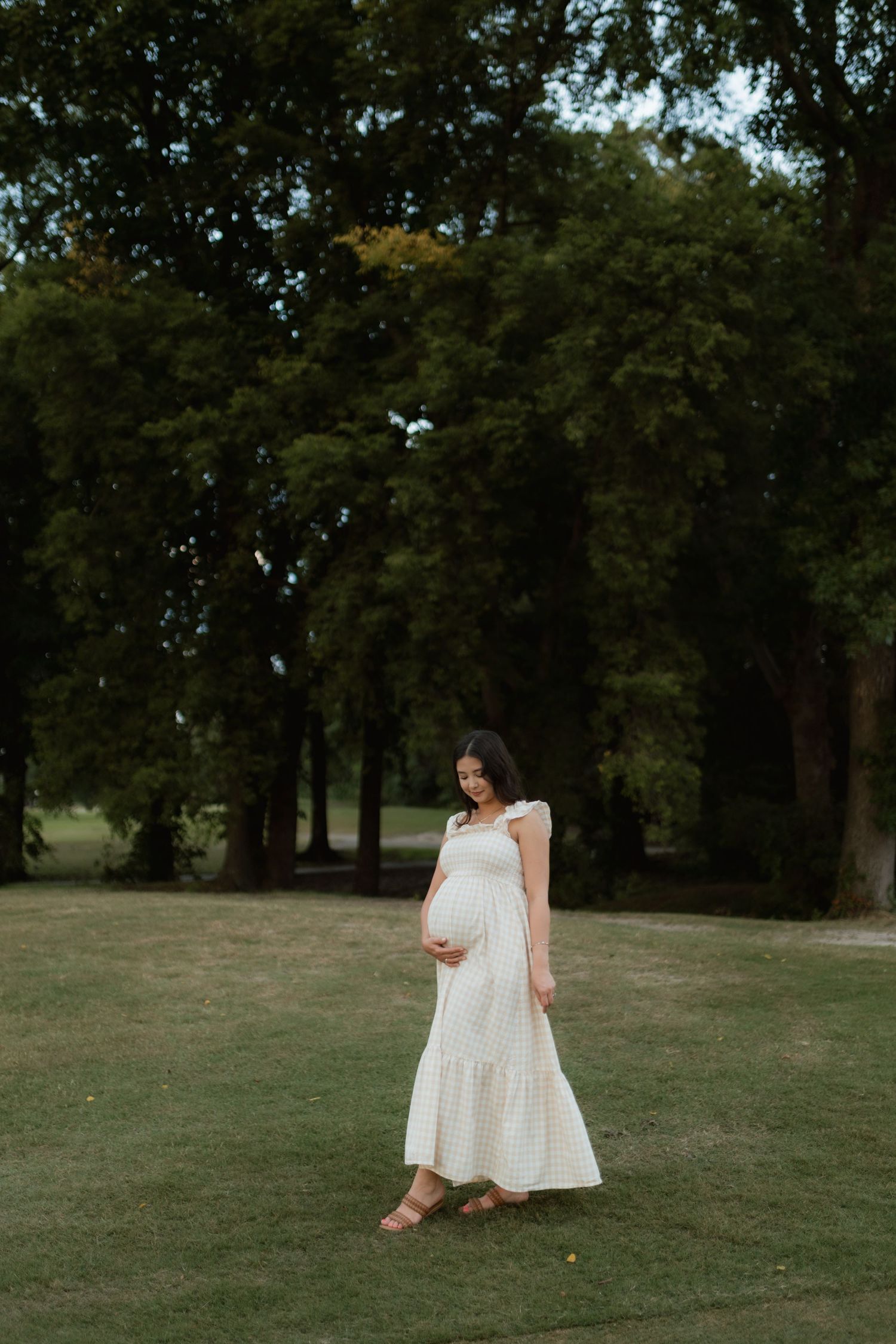Pregnant woman in white maxi dress standing in green park, gently holding her baby bump.