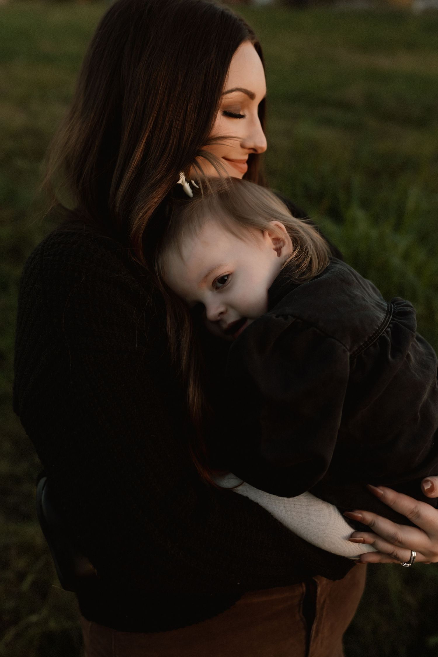 Mother with dark hair tenderly holds sleeping toddler in black outfit at golden hour.