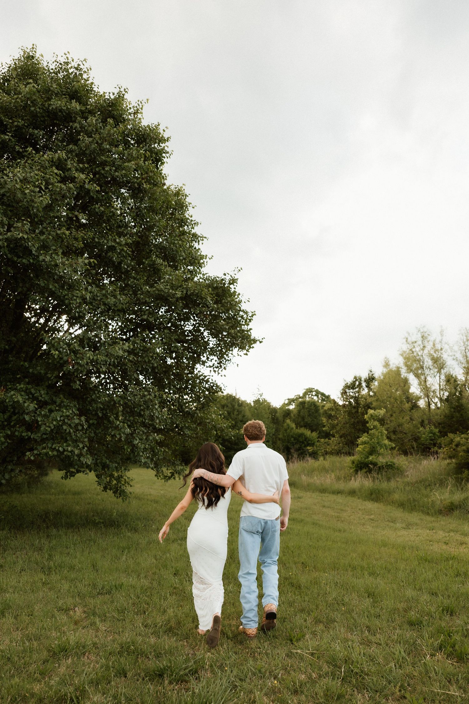A couple walks arm in arm through a green meadow with trees under a cloudy sky.