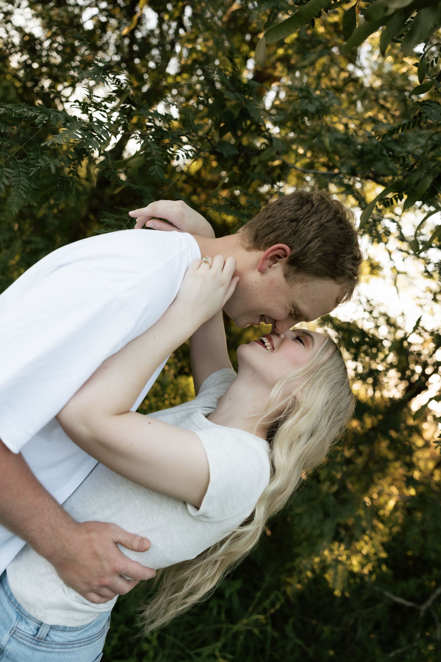 A loving couple sharing a kiss while standing close together near lush green foliage in warm sunlight.