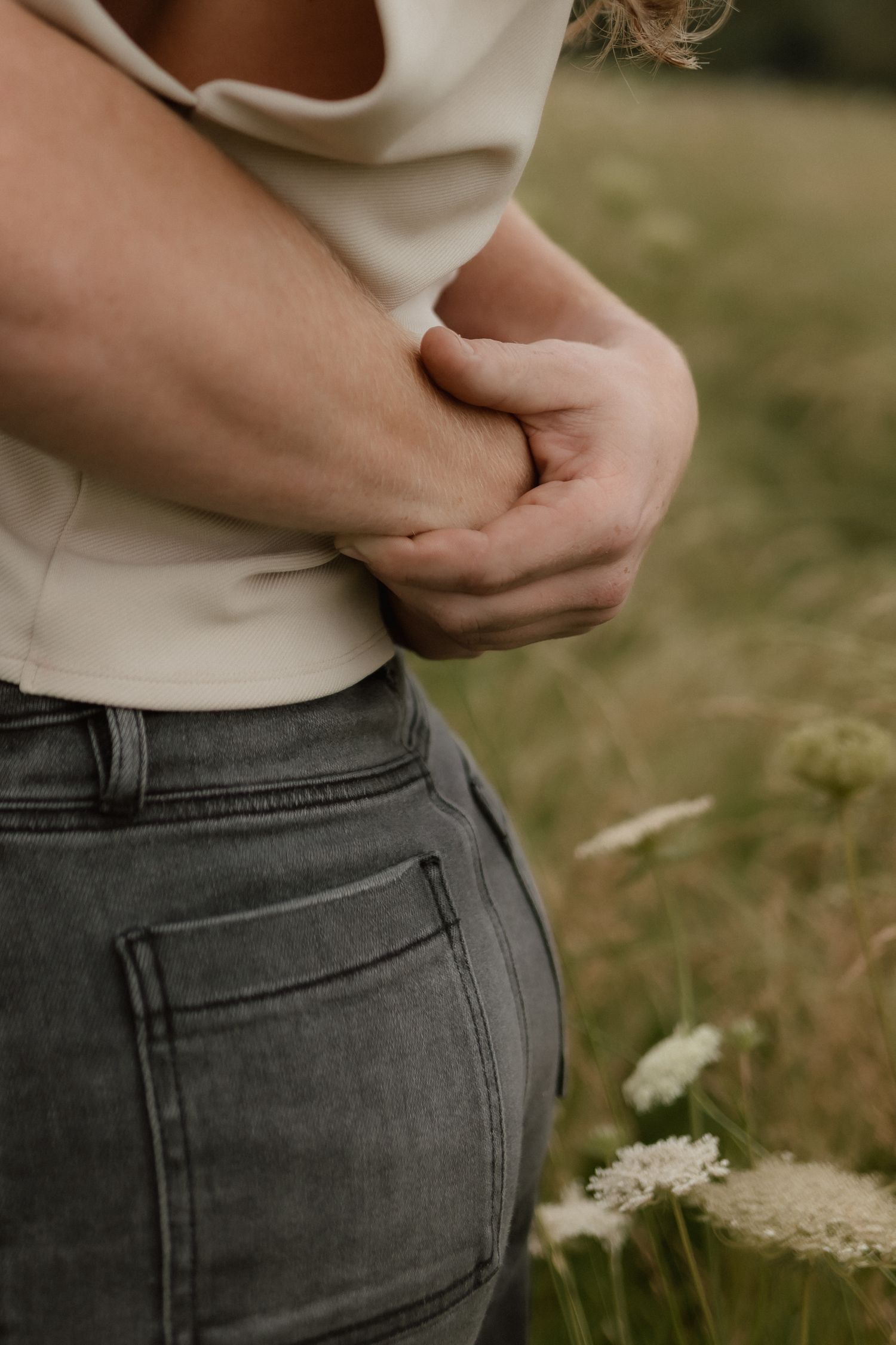 Close-up of a couple holding hands near waist, woman wearing gray jeans and beige top in a grassy field.