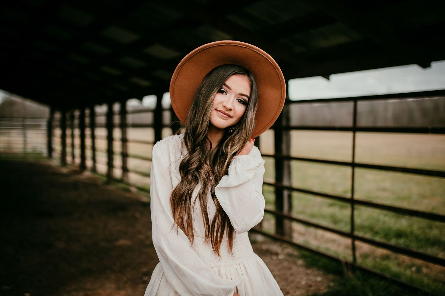 Brunette woman in wide-brim tan hat and white dress poses softly beside a rustic outdoor fence.
