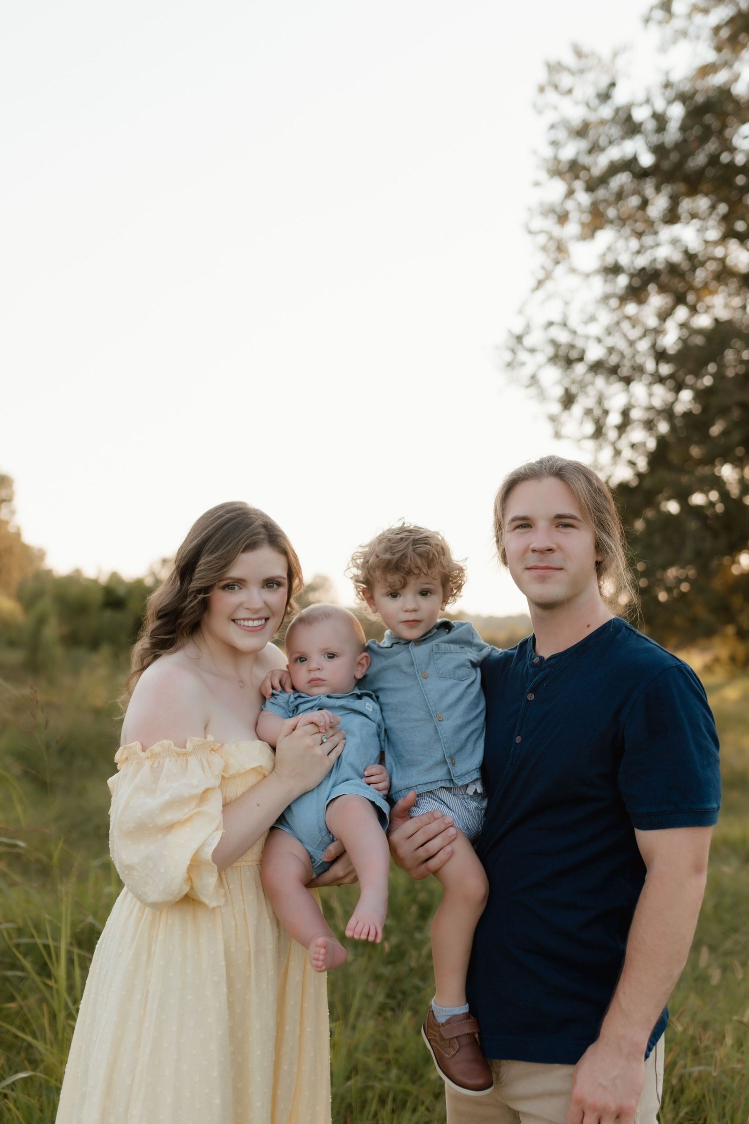Happy family portrait in sunlit meadow, mother in yellow dress holding infant beside father and curly-haired toddler.