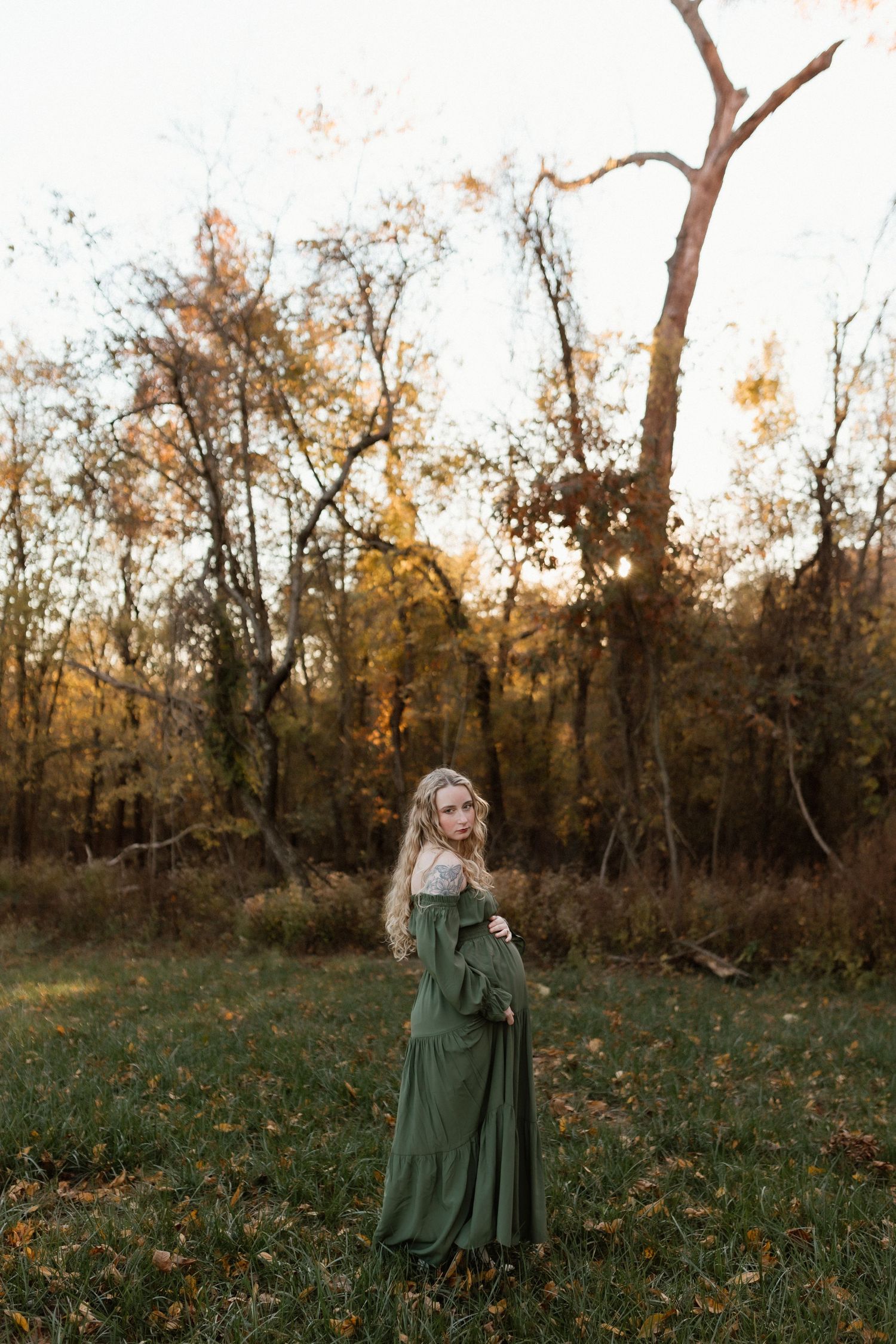 Pregnant woman in elegant green maxi dress stands in fall woodland during golden hour.