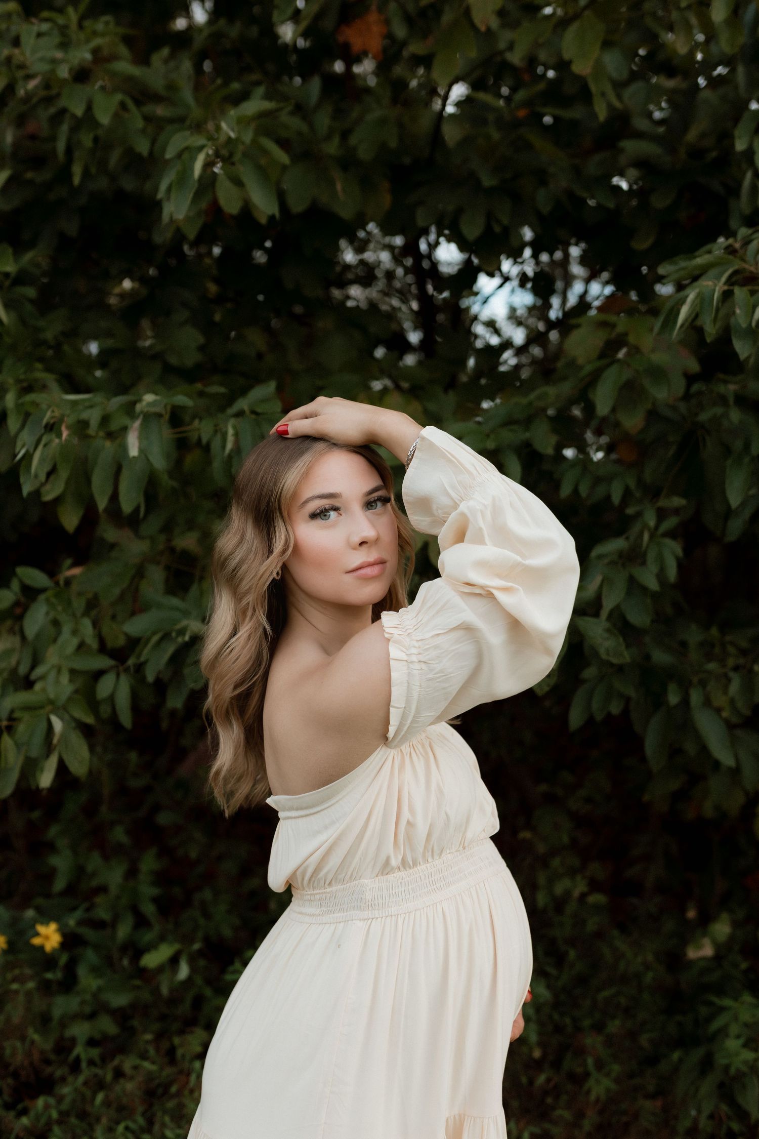 Pregnant woman in cream off-shoulder dress poses gracefully among lush green trees.