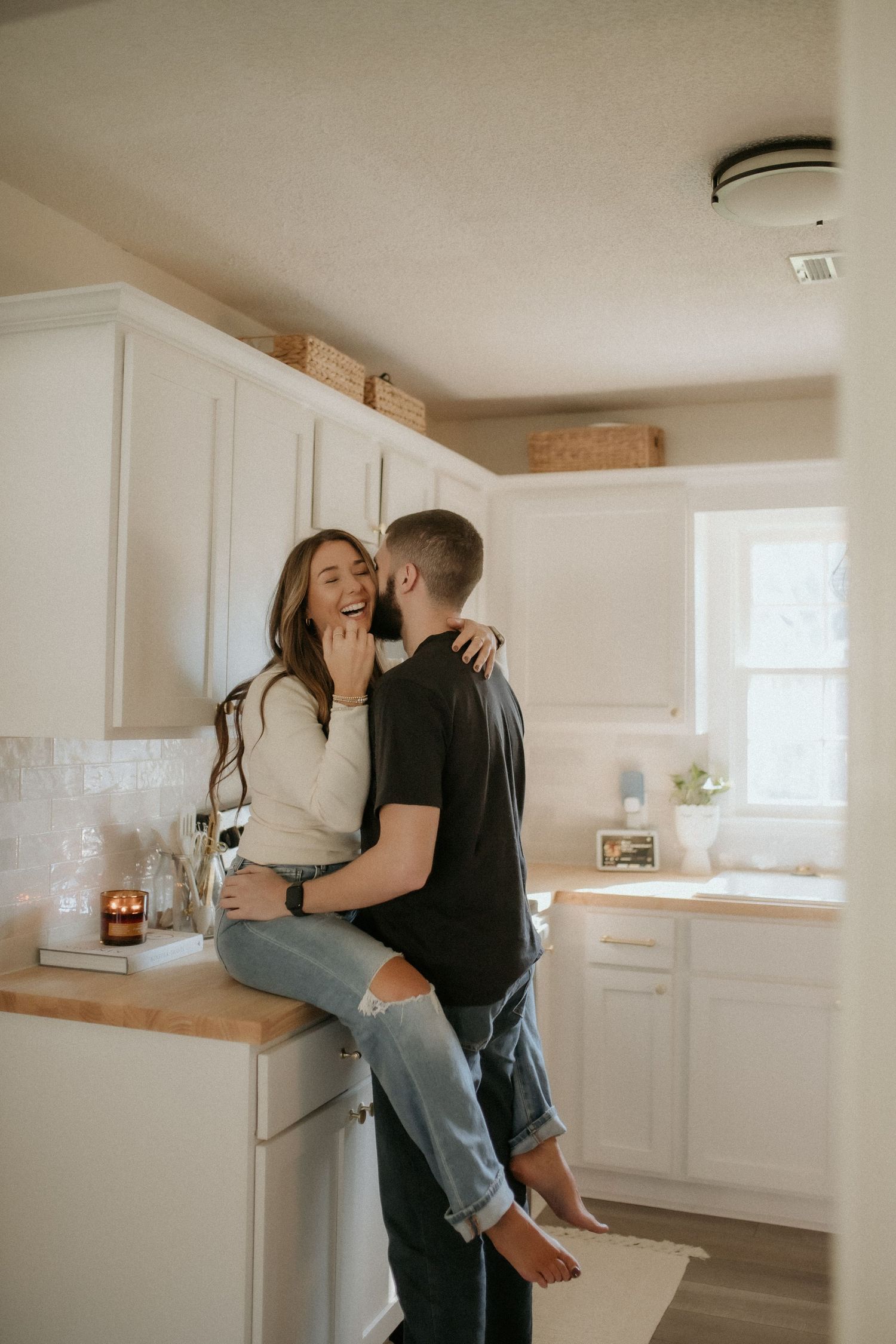 Laughing brunette woman sits on kitchen counter embracing her partner in a bright white cozy home.