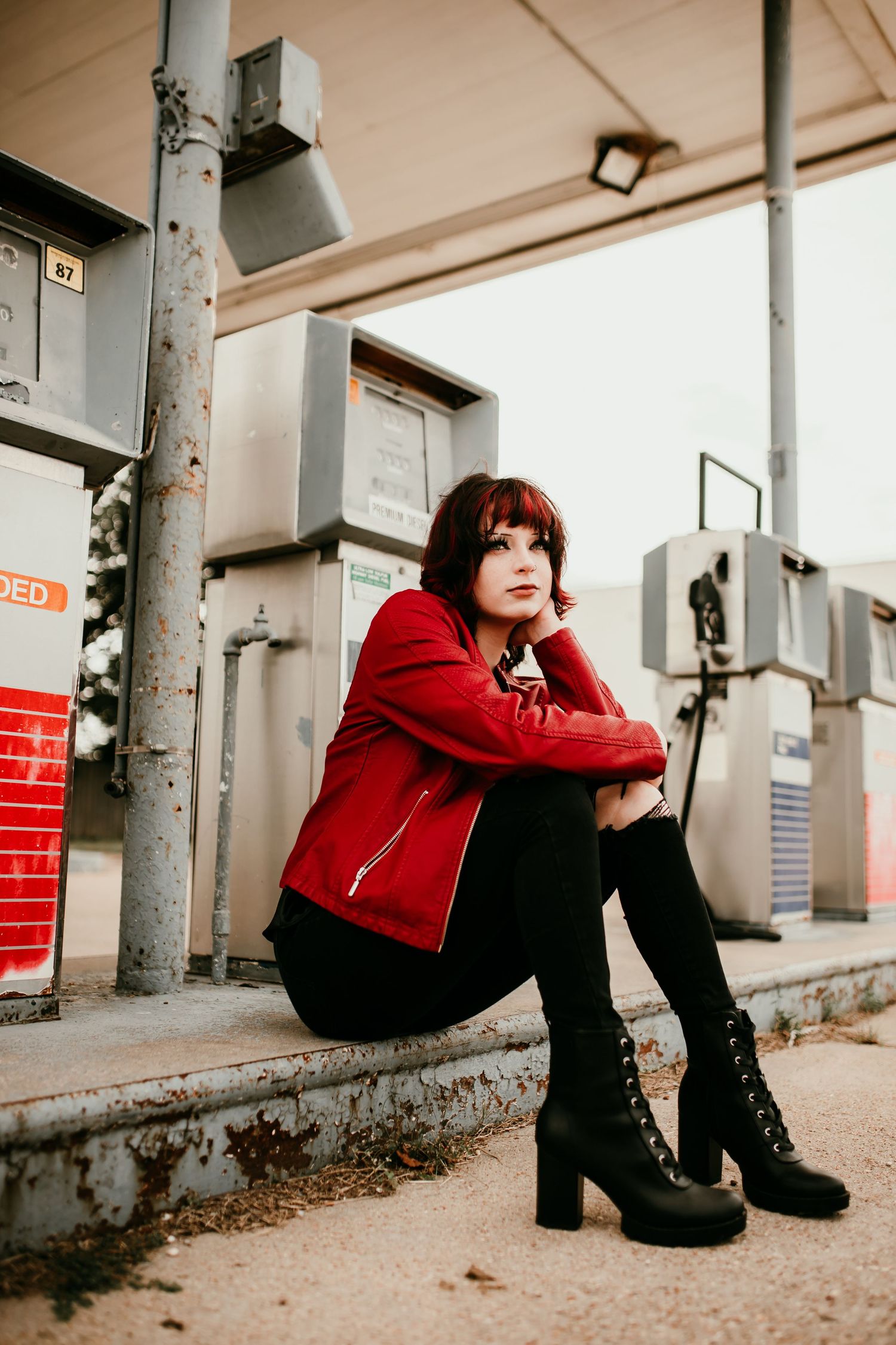 Young woman with red hair in red leather jacket and black boots sitting at an abandoned gas station.