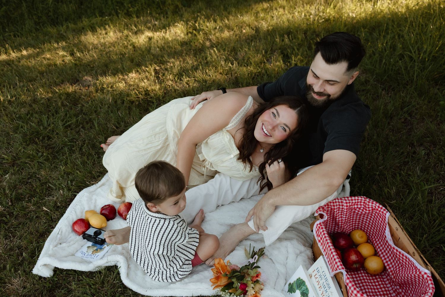 Family enjoying a picnic on a white blanket in the grass with fresh fruits and a red gingham basket.