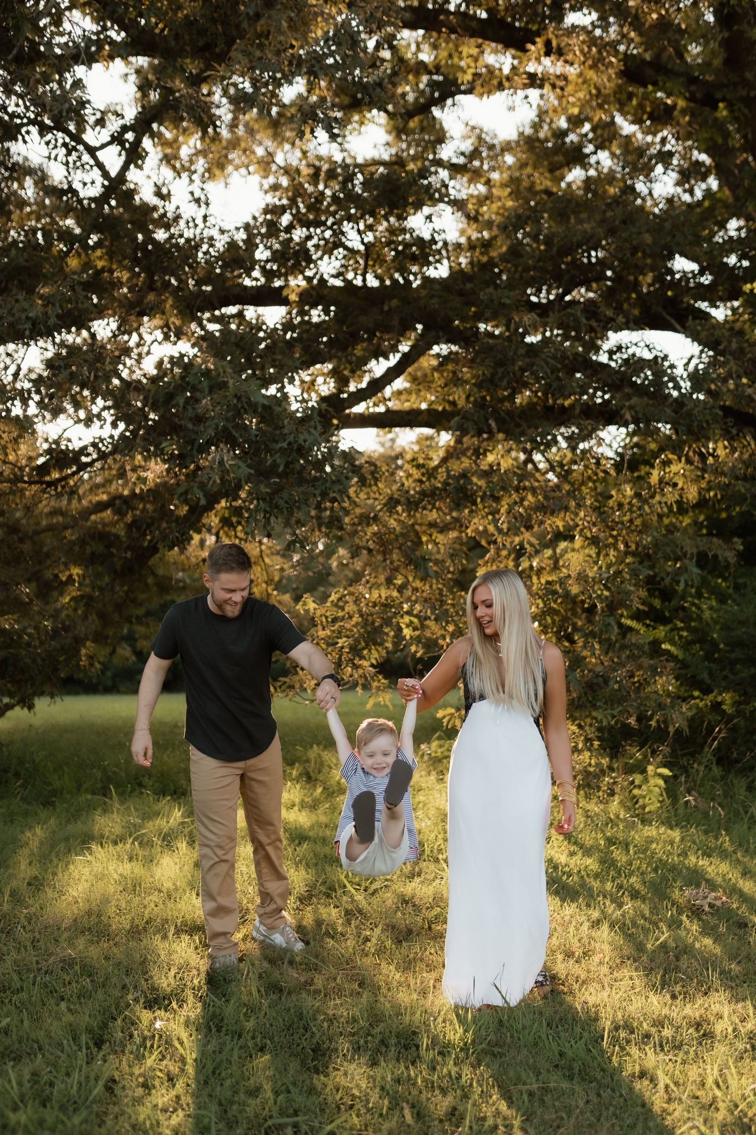 Family swinging young child between parents under golden sunlit tree in grassy field.