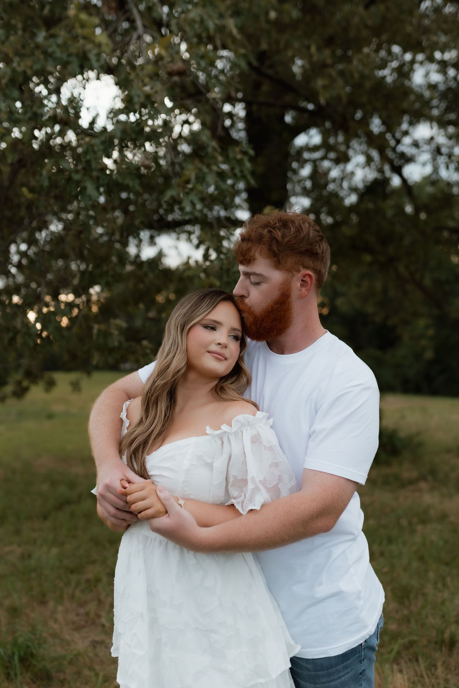 A man with red hair kisses a woman on the head while embracing her in a white dress outdoors.