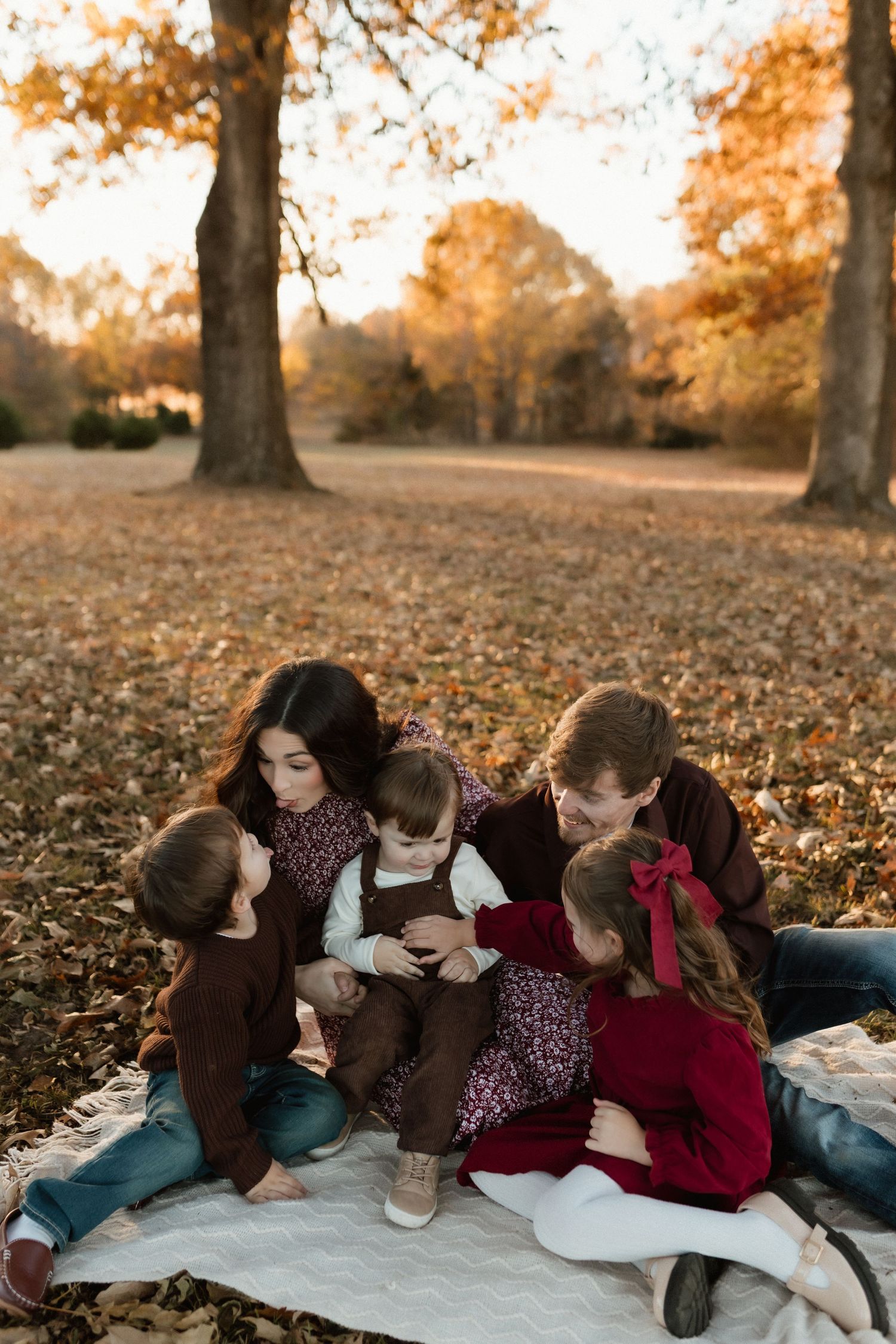 Family of five sits together on a blanket in a leaf-covered park, sharing a tender moment at golden hour.