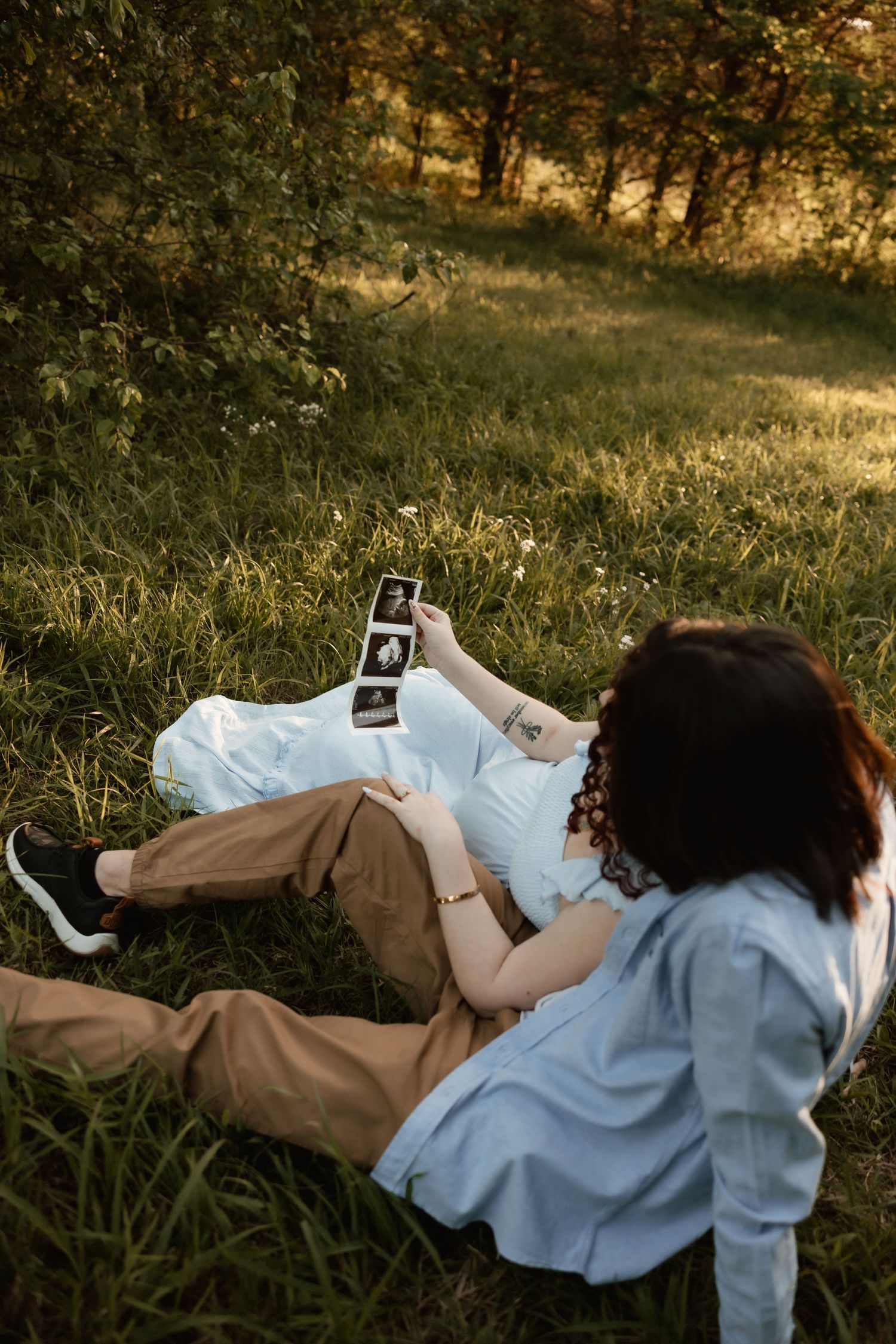 Pregnant couple sitting in golden sunlit grass reviewing ultrasound photos together outdoors.