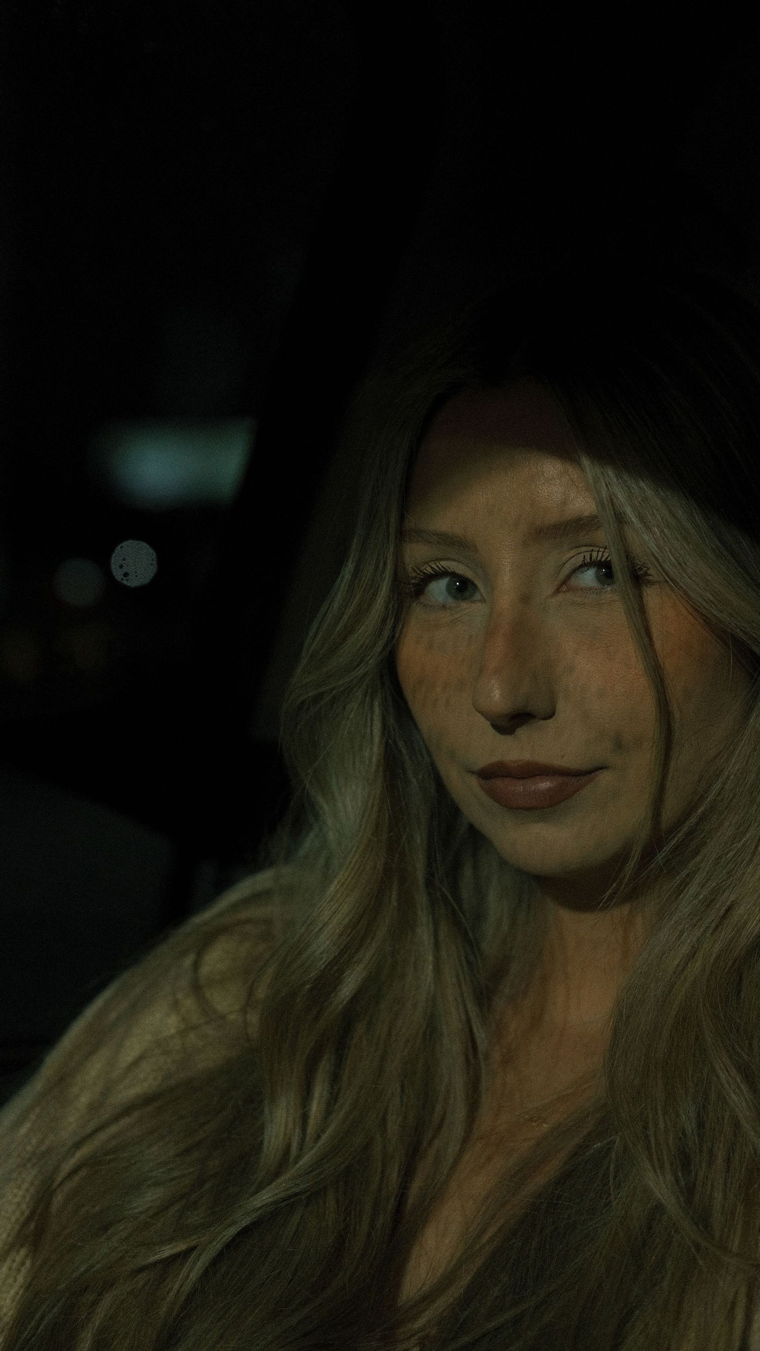 Young woman with long blonde hair poses in a dimly lit car at night with bokeh lights in background.