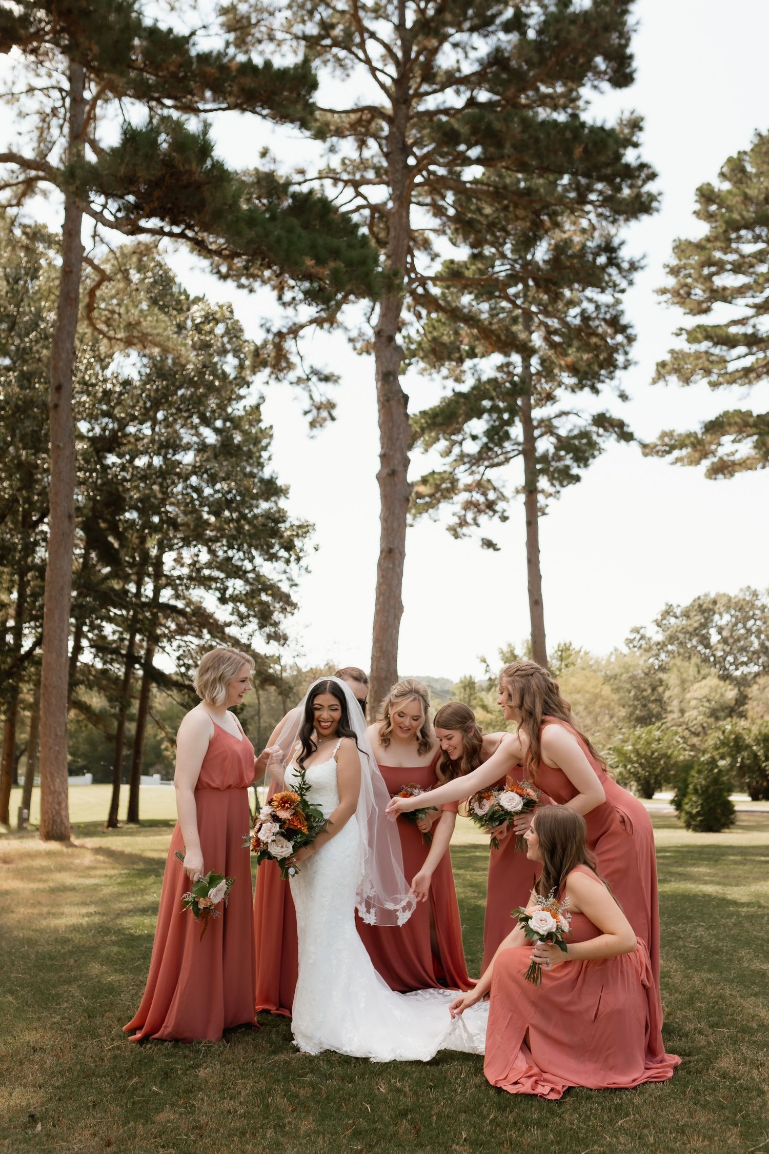 Bride surrounded by bridesmaids in rust-colored gowns holding bouquets beneath tall pine trees.