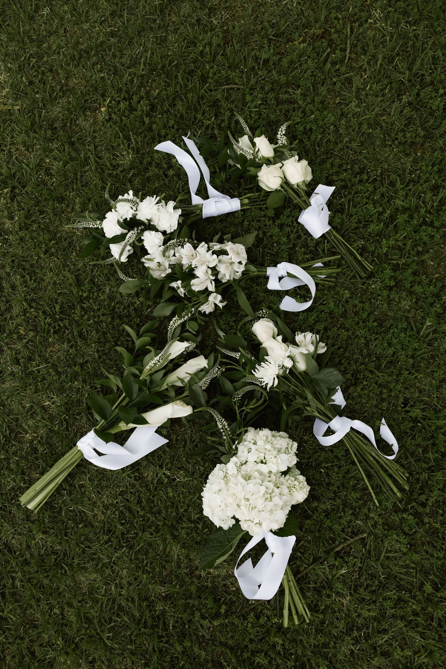 Four white wedding bouquets with ribbon ties arranged on green grass at outdoor wedding.