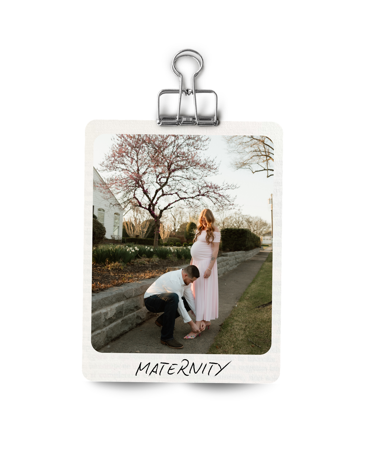Maternity photo displayed as a Polaroid clip showing couple outdoors near blooming spring trees on a path.