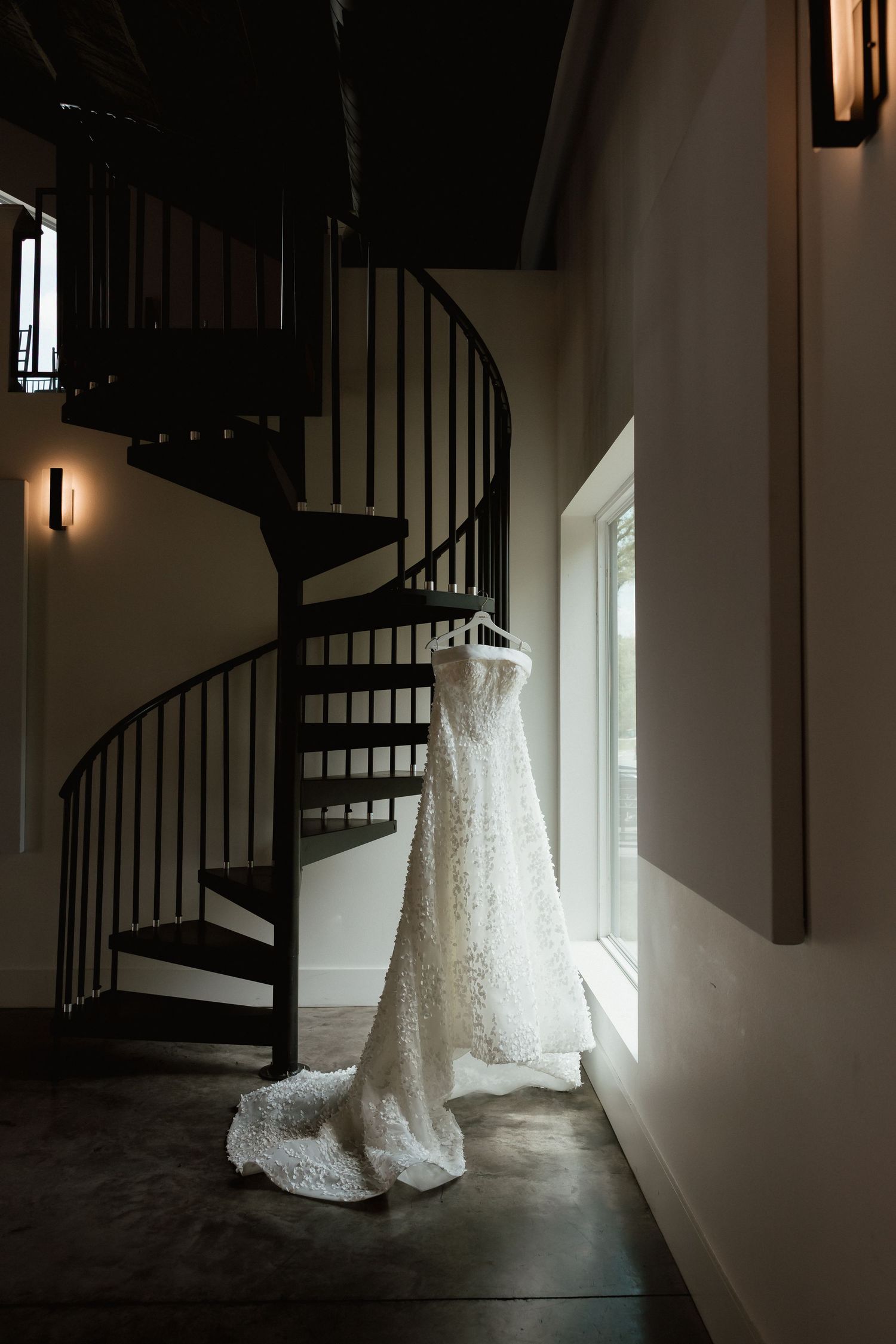 Elegant lace wedding dress hanging near spiral staircase in beautiful natural window light.