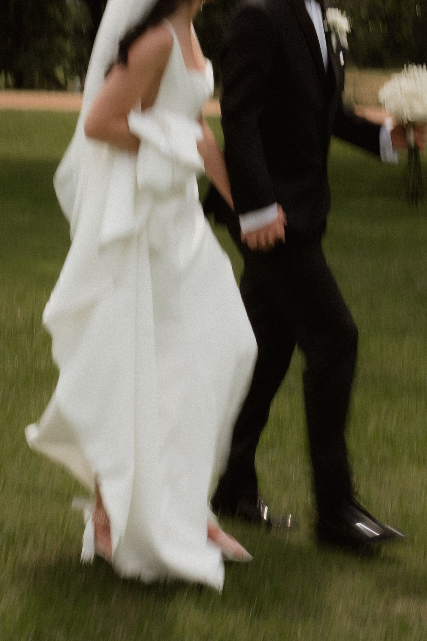 Motion blur captures bride in white gown and groom in black tuxedo walking together on grass.
