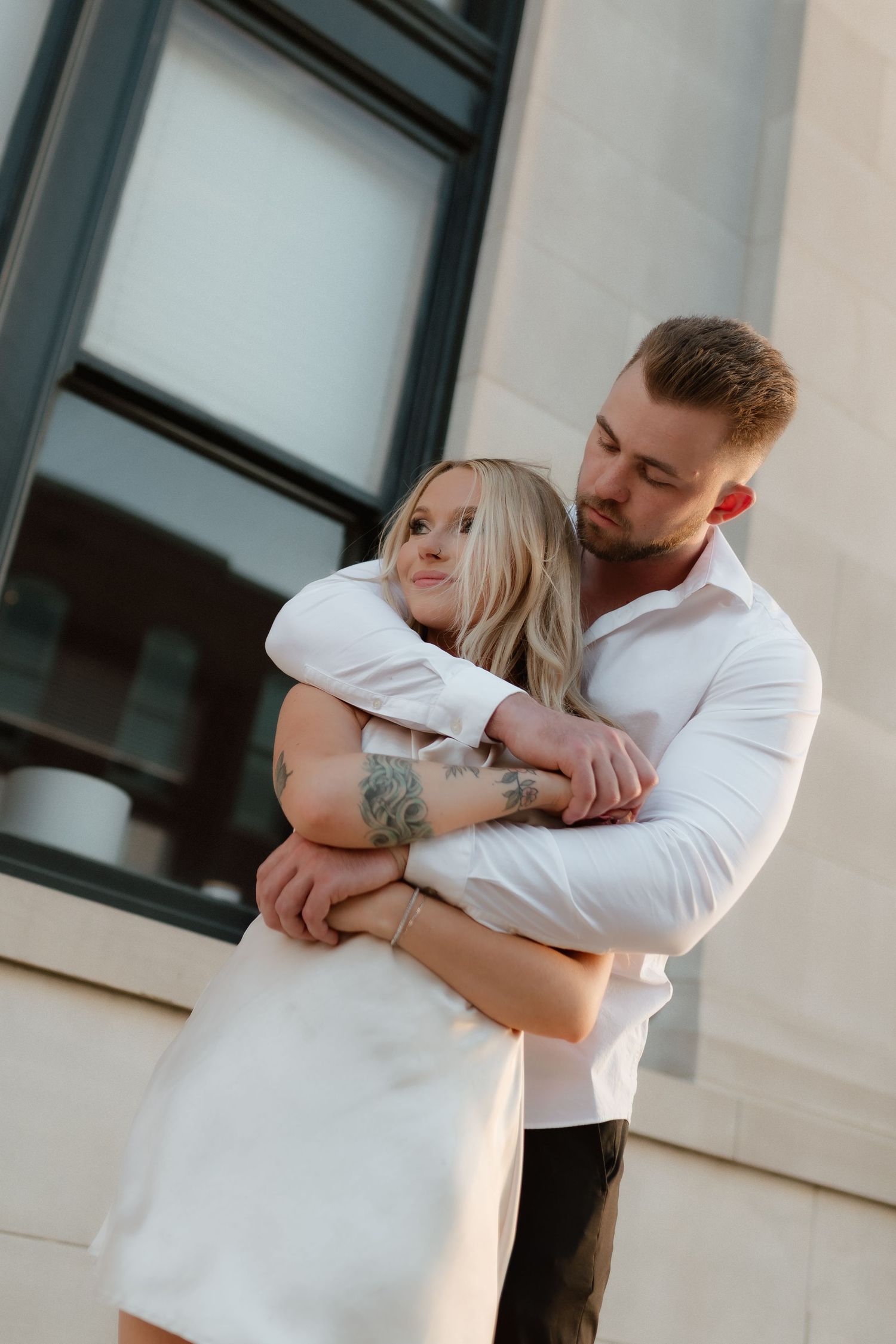 A tattooed couple shares an intimate embrace against a stone building exterior at dusk.