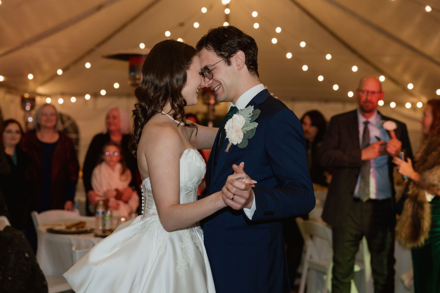 Bride and groom sharing first dance under warm string lights surrounded by wedding guests.