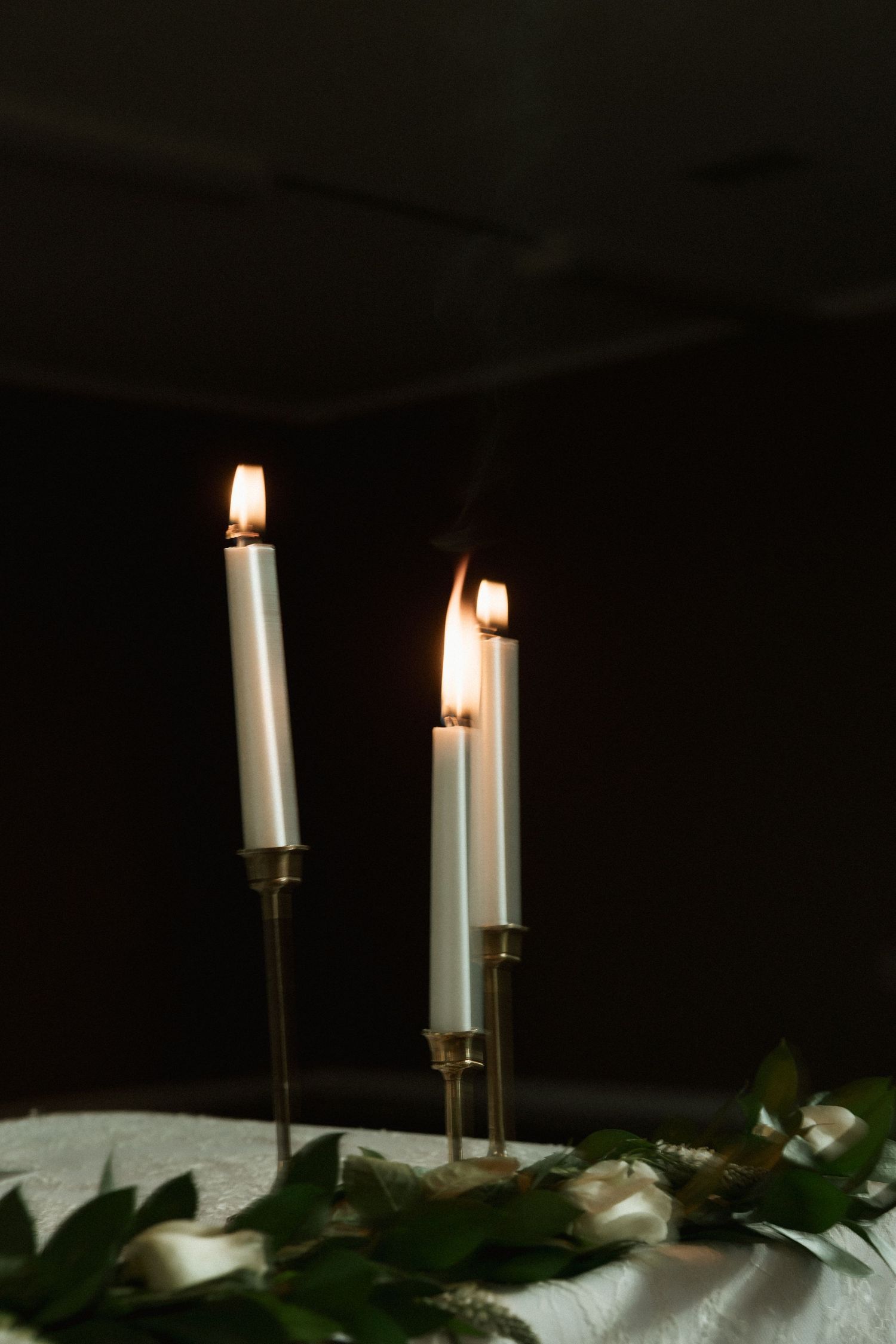 Three lit white taper candles in gold holders on a unity candle table at wedding ceremony.