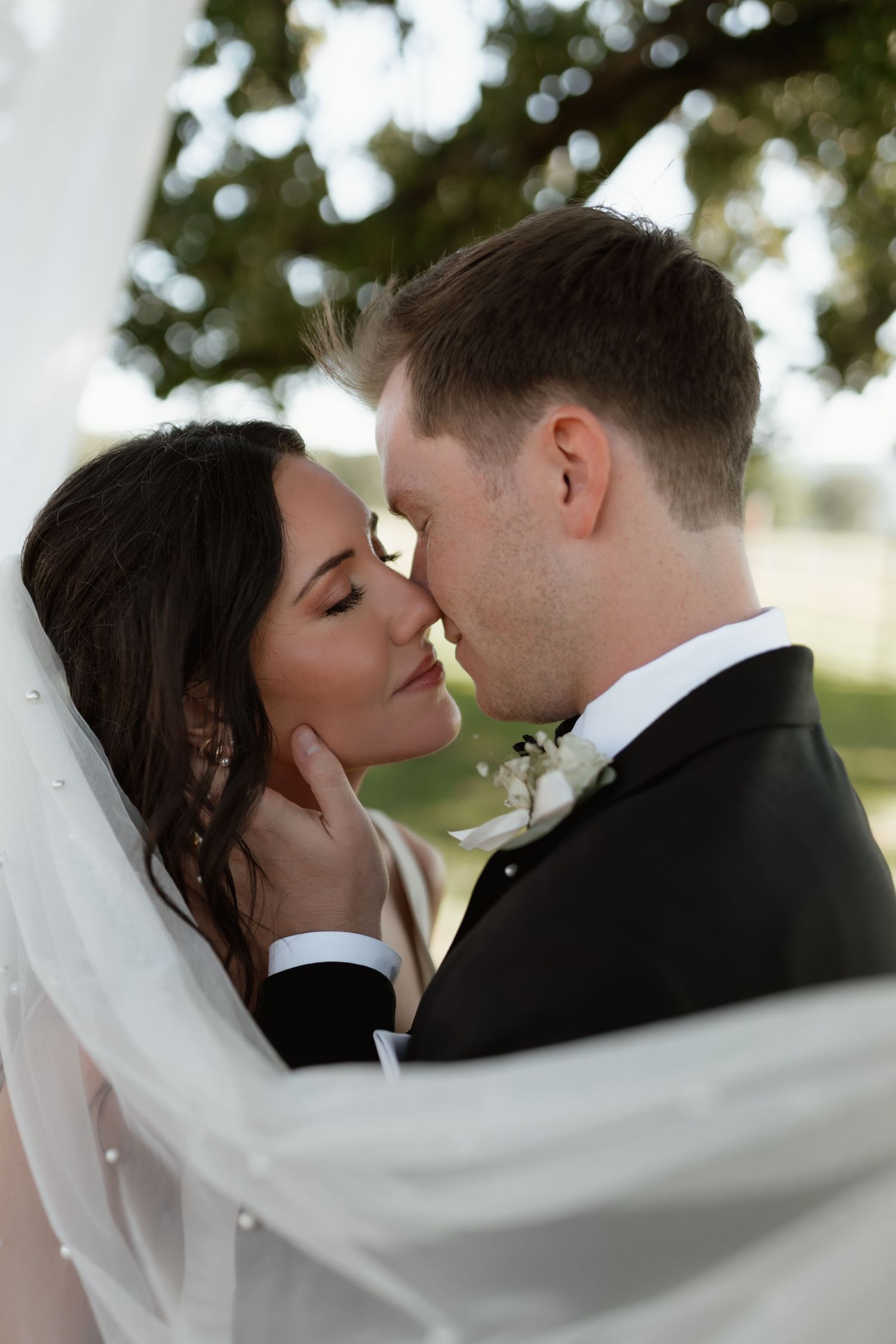Groom tenderly cups brides face as they share an intimate moment framed by a flowing white veil.