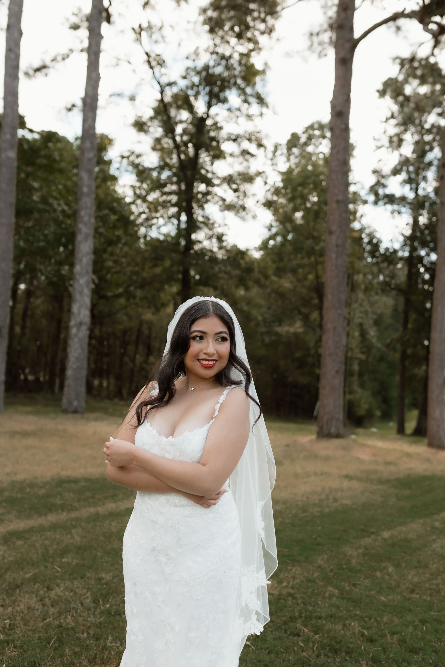 Smiling bride in a lace wedding gown and veil posing outdoors among tall trees on her wedding day.