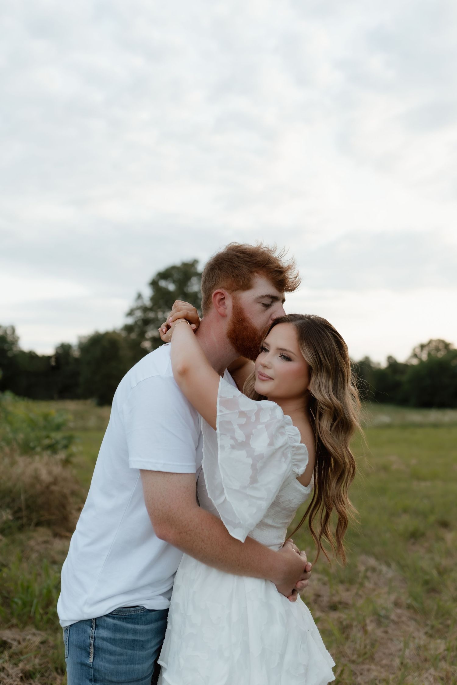 A bearded man in a white shirt embraces a woman in a white lace dress in an open field.