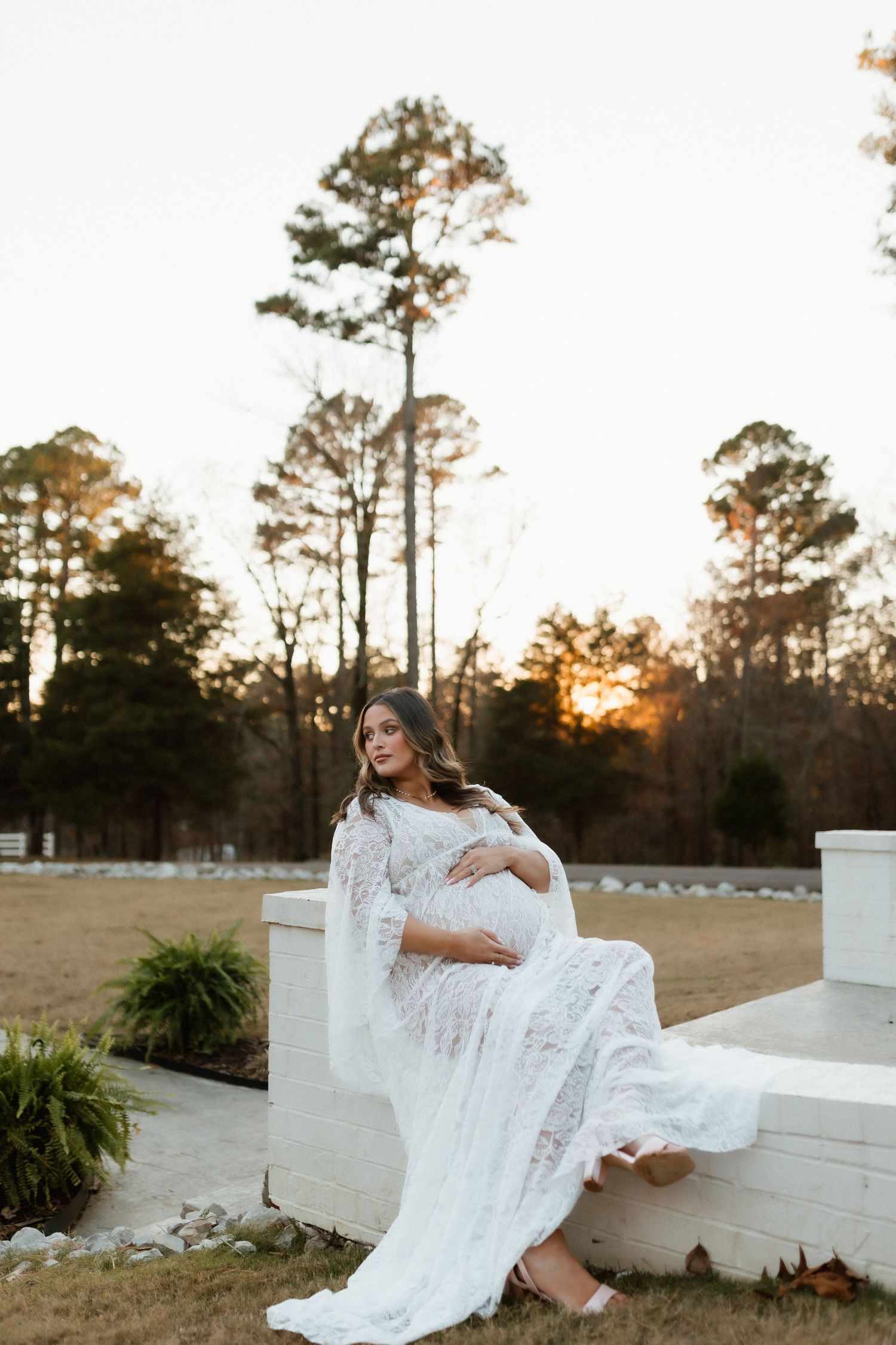 Pregnant woman in flowing white lace gown sitting on stone steps at sunset, cradling baby bump.