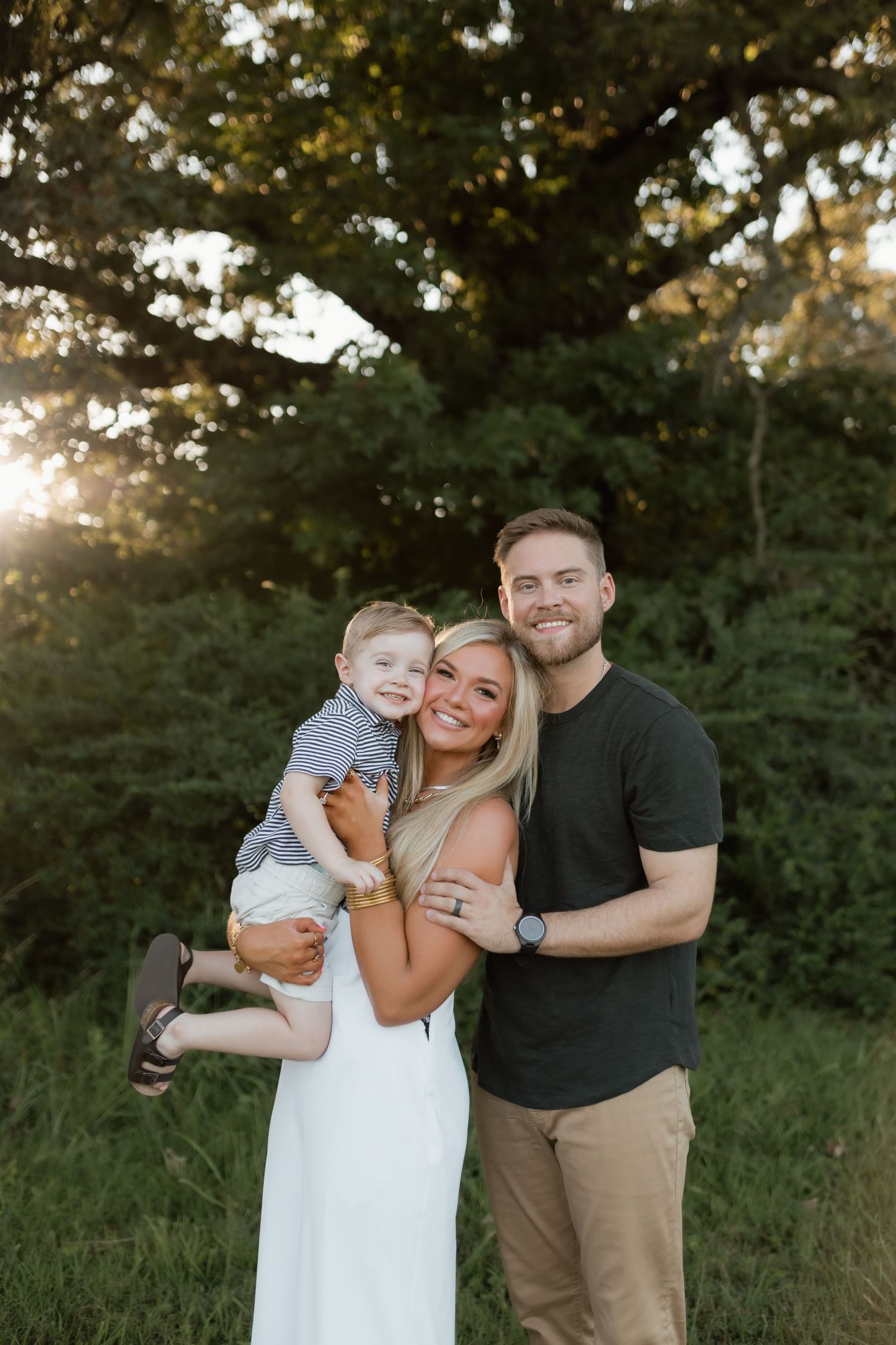 Smiling family with blonde mother holding toddler and father posing in sunlit green field.