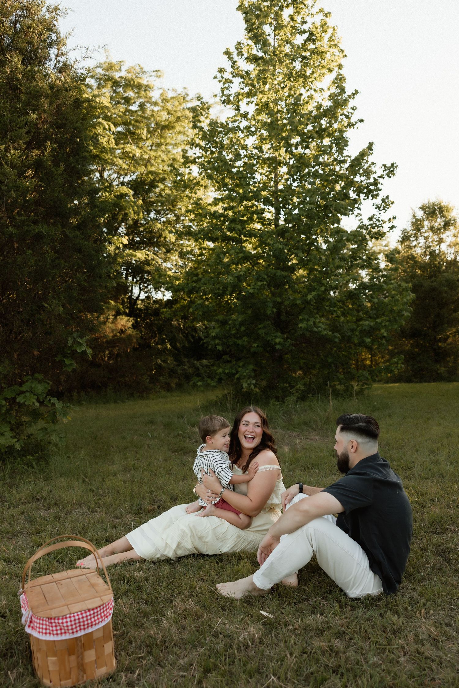 Happy family with baby sitting on grass during outdoor picnic with wicker basket and trees.