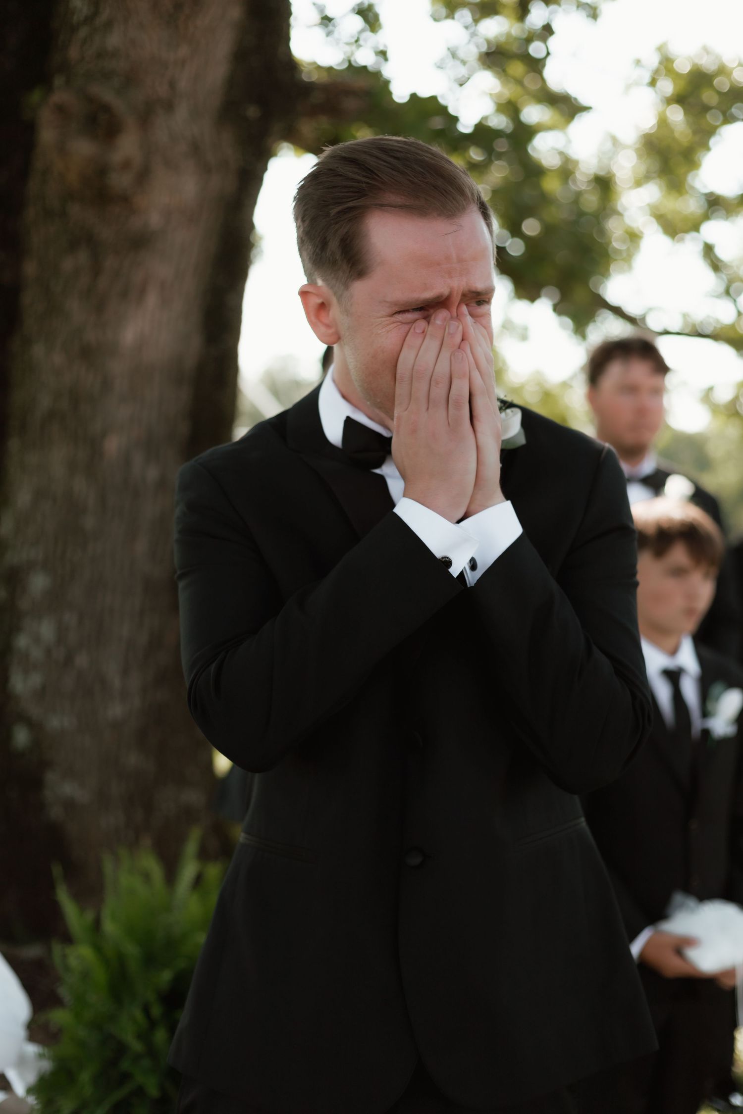 Tearful groom in black tuxedo covers face with hands at outdoor wedding ceremony.
