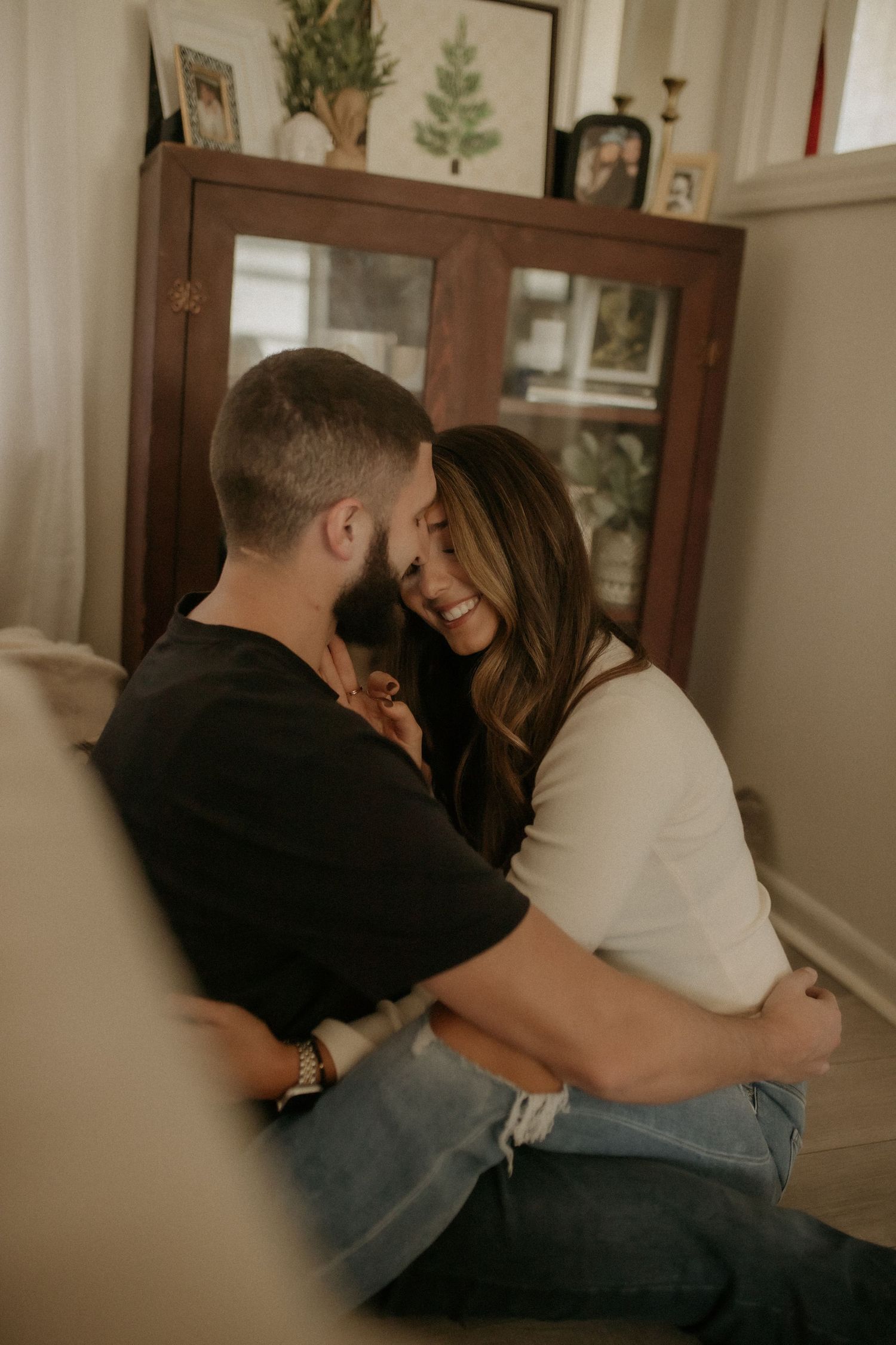 Couple embraces and smiles closely together while sitting on a couch in a warmly decorated living room.